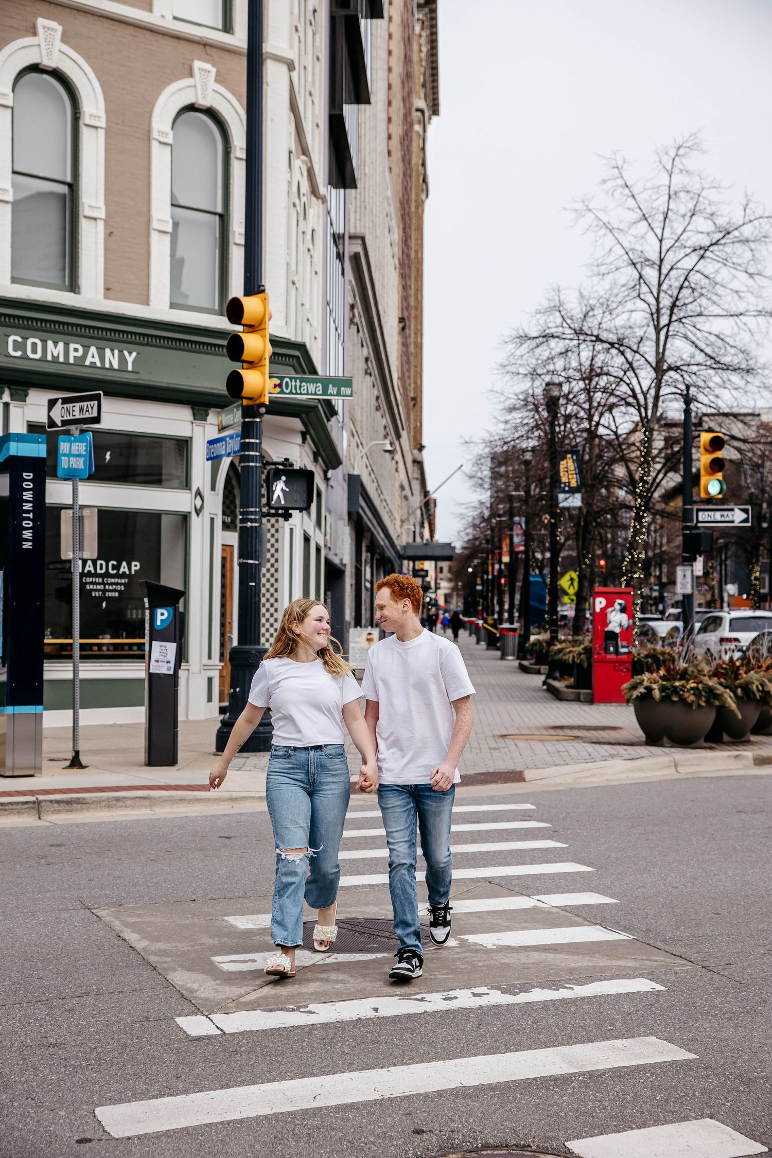 A couple walks together holding hands while crossing a city street crosswalk on a cloudy urban day.
