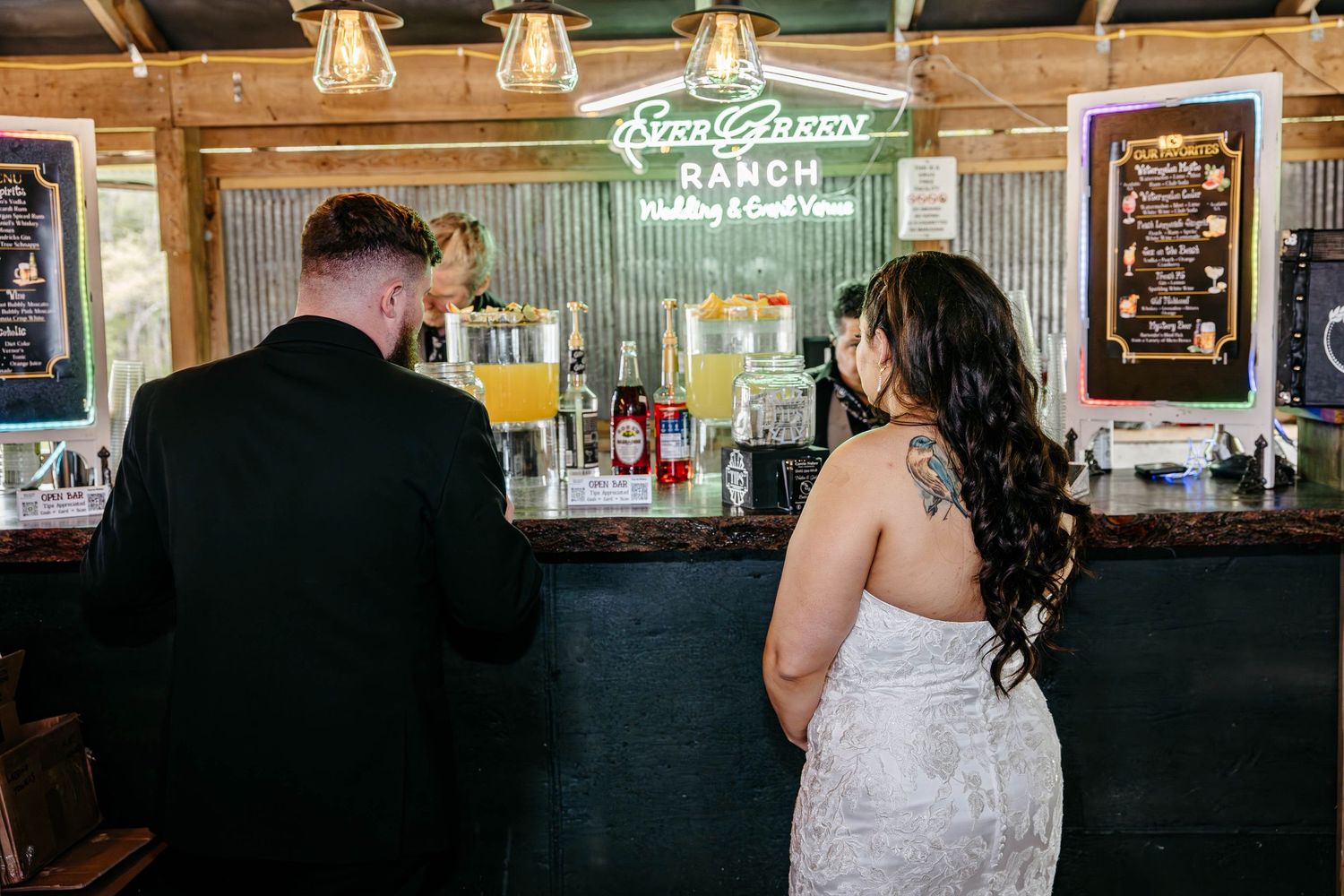 A couple sits at a rustic wooden bar together with illuminated signage glowing green behind them.