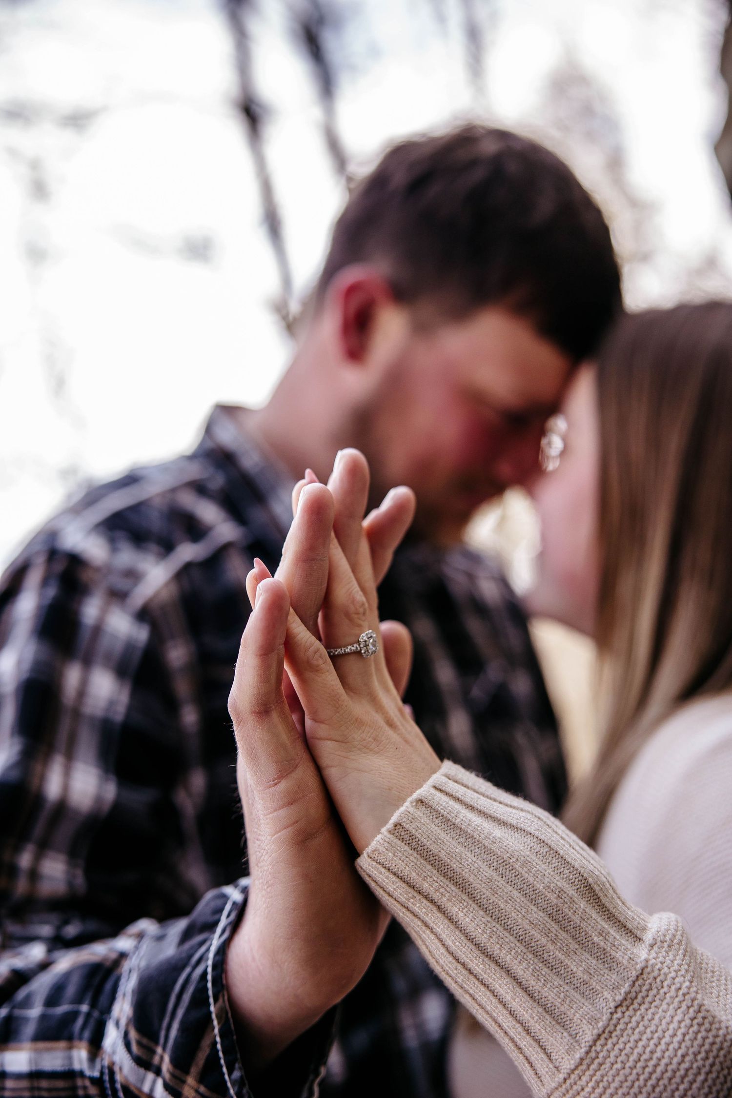 Close-up of hands with engagement ring showing, with a couple embracing in plaid shirt and sweater in soft focus background.
