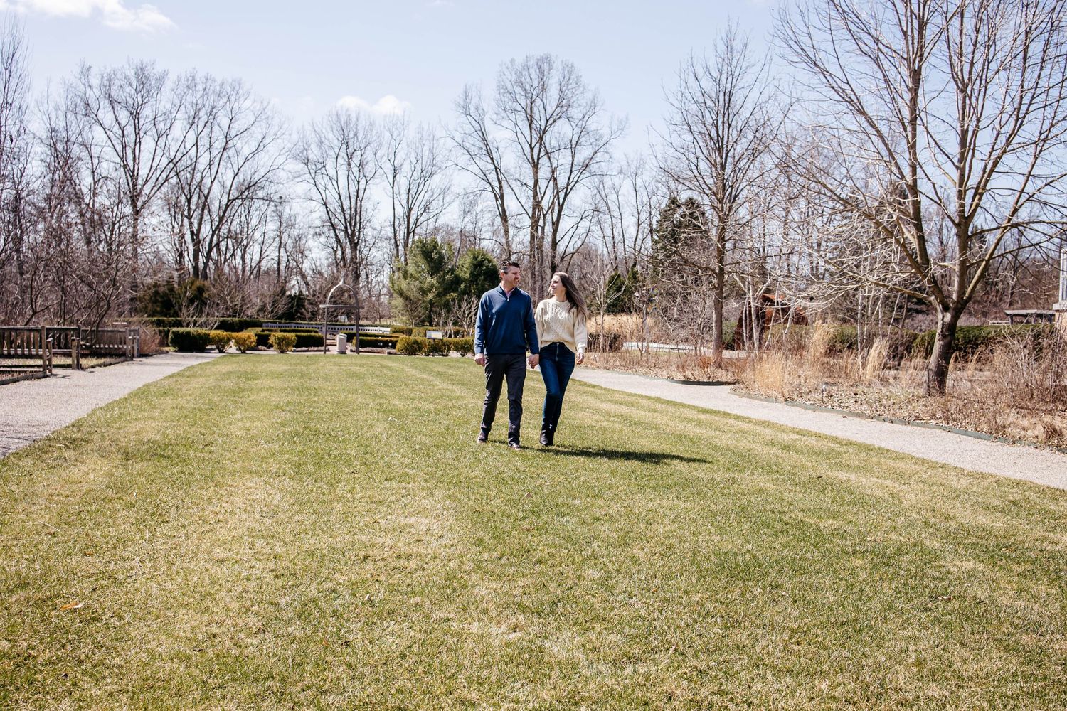 A couple walks together on a grassy path surrounded by bare trees on a sunny winter day.