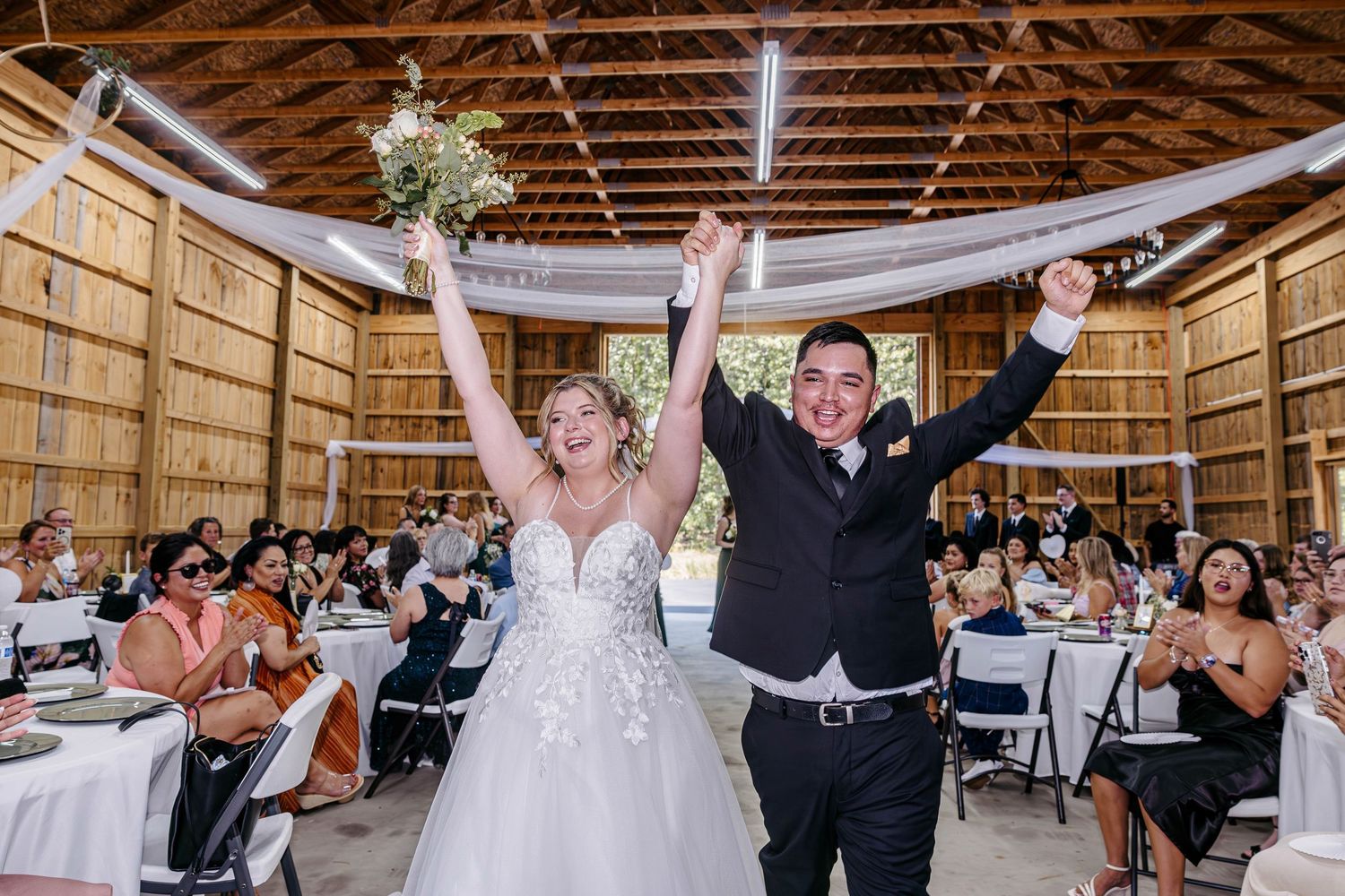 A joyful wedding celebration inside a rustic wooden barn decorated with white draping and filled with seated guests.