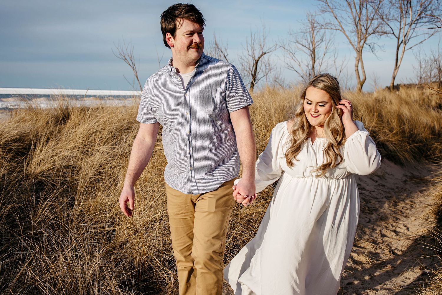 A couple walks hand in hand along a beach path through tall grass on a sunny day.