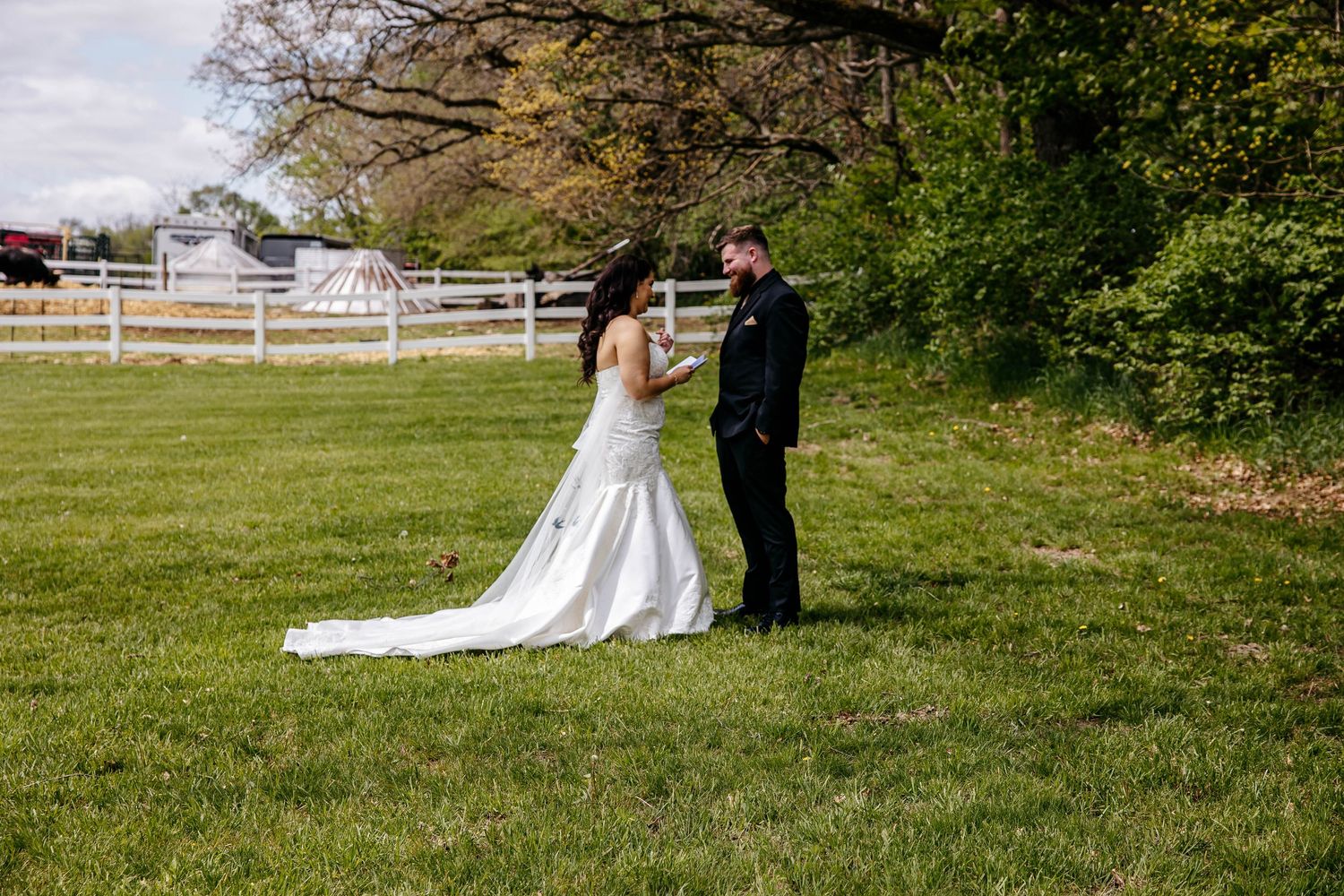 A couple in wedding attire stands together on a green lawn with a white fence and trees in the background.