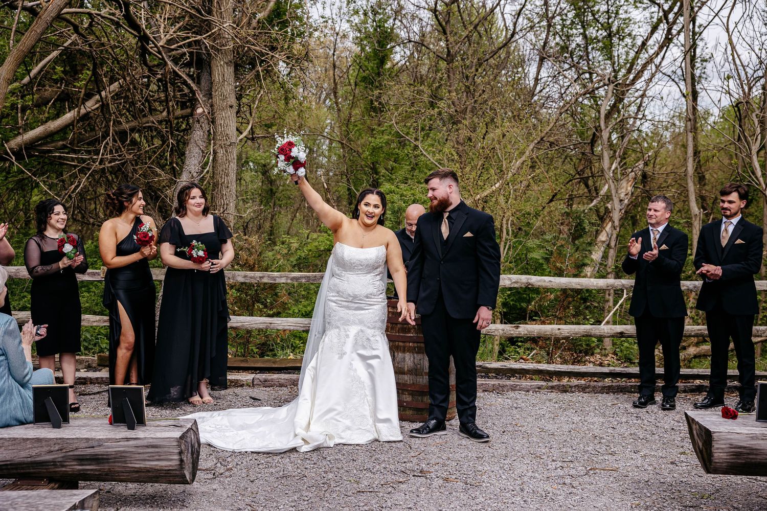 A wedding ceremony takes place outdoors on a gravel path with a wooden fence and trees in the background.
