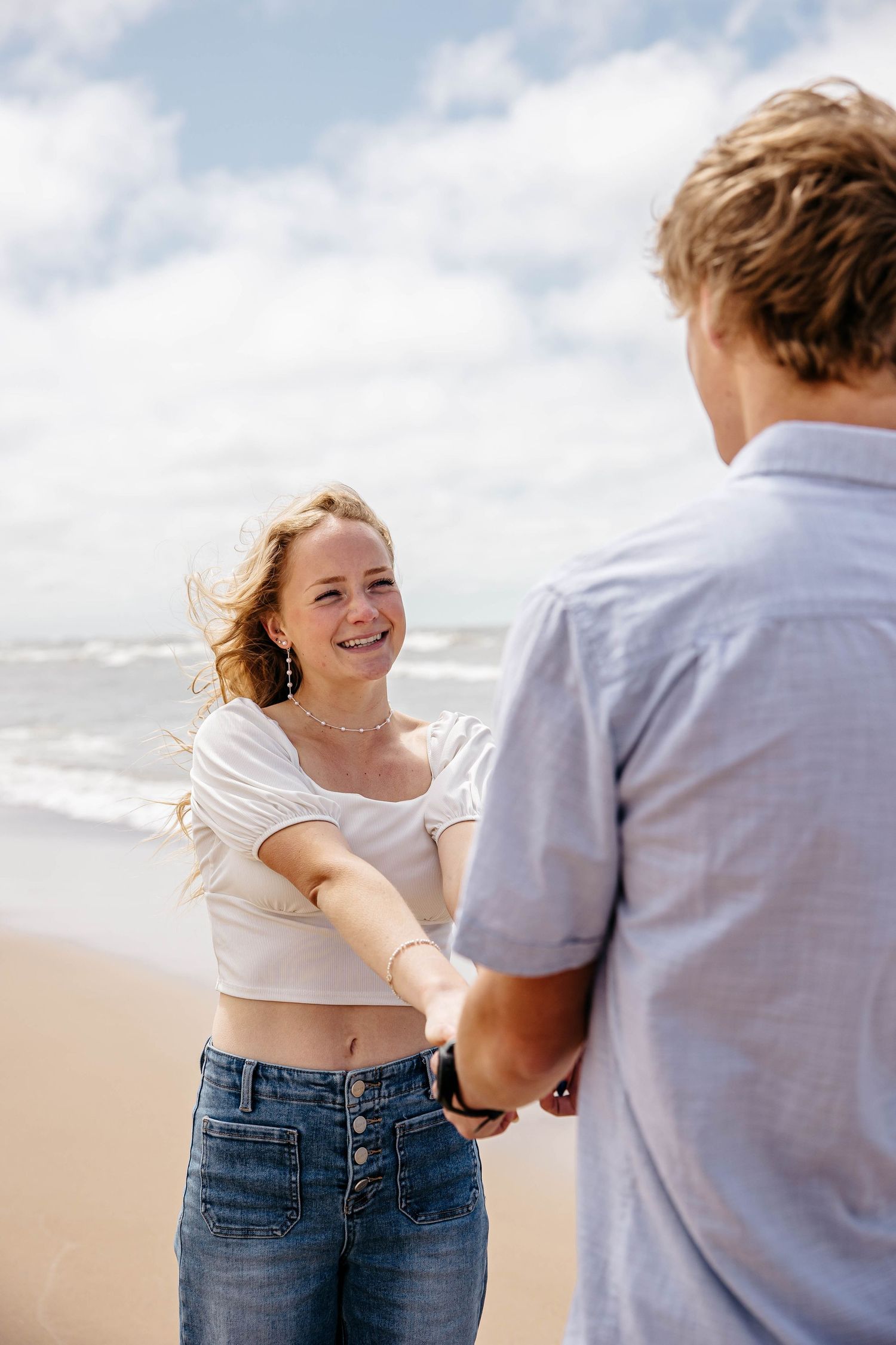 Couple holding hands and sharing a romantic moment together on a sunny beach with waves and blue sky in background.