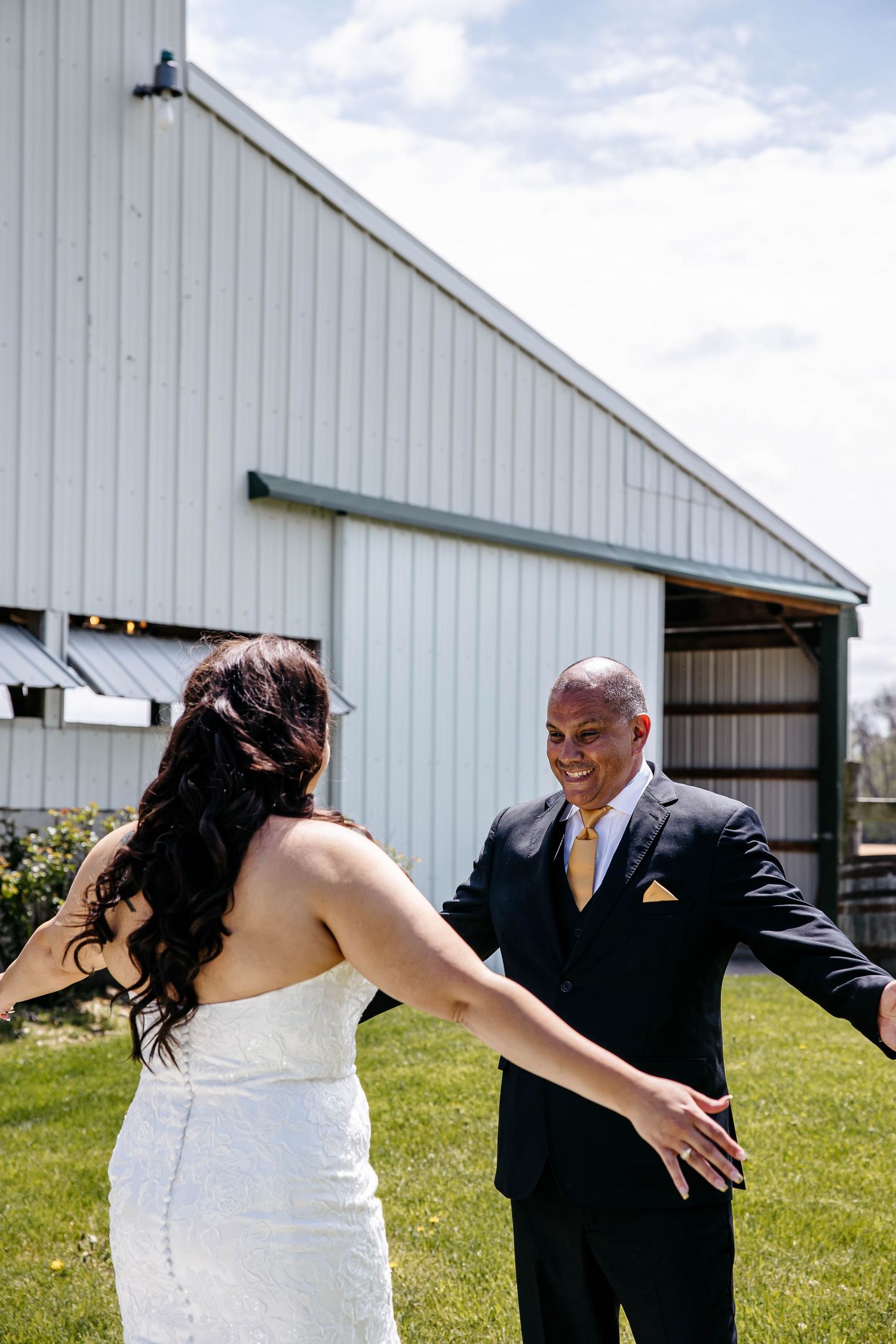 Newlyweds share a playful first look moment outdoors with white barns in the background on their wedding day.