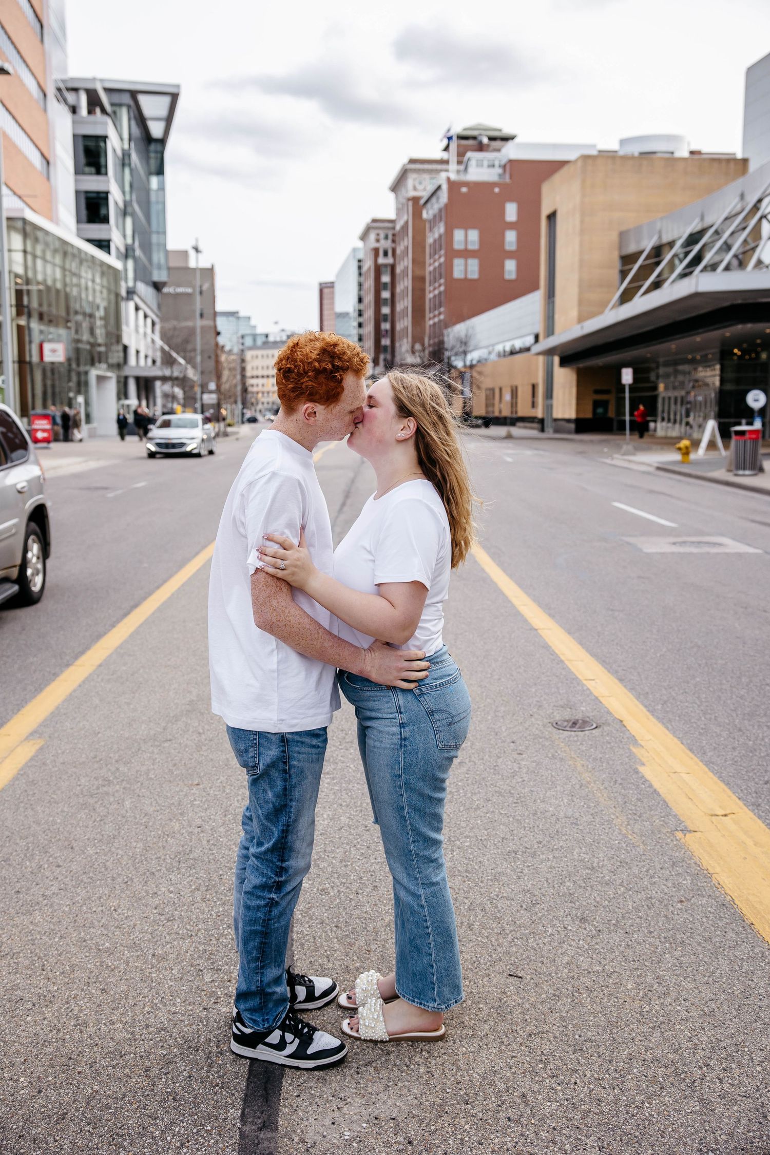 Young couple in white tops and jeans share a romantic kiss while standing in the middle of a city street.