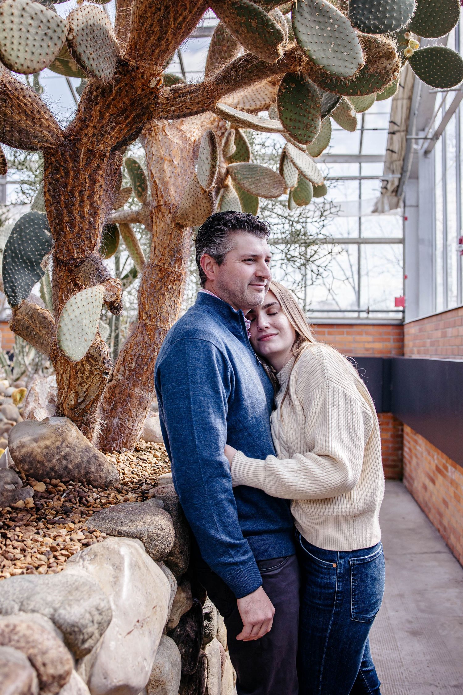 A couple embraces tenderly in a greenhouse setting with large cacti and natural light streaming through the windows.