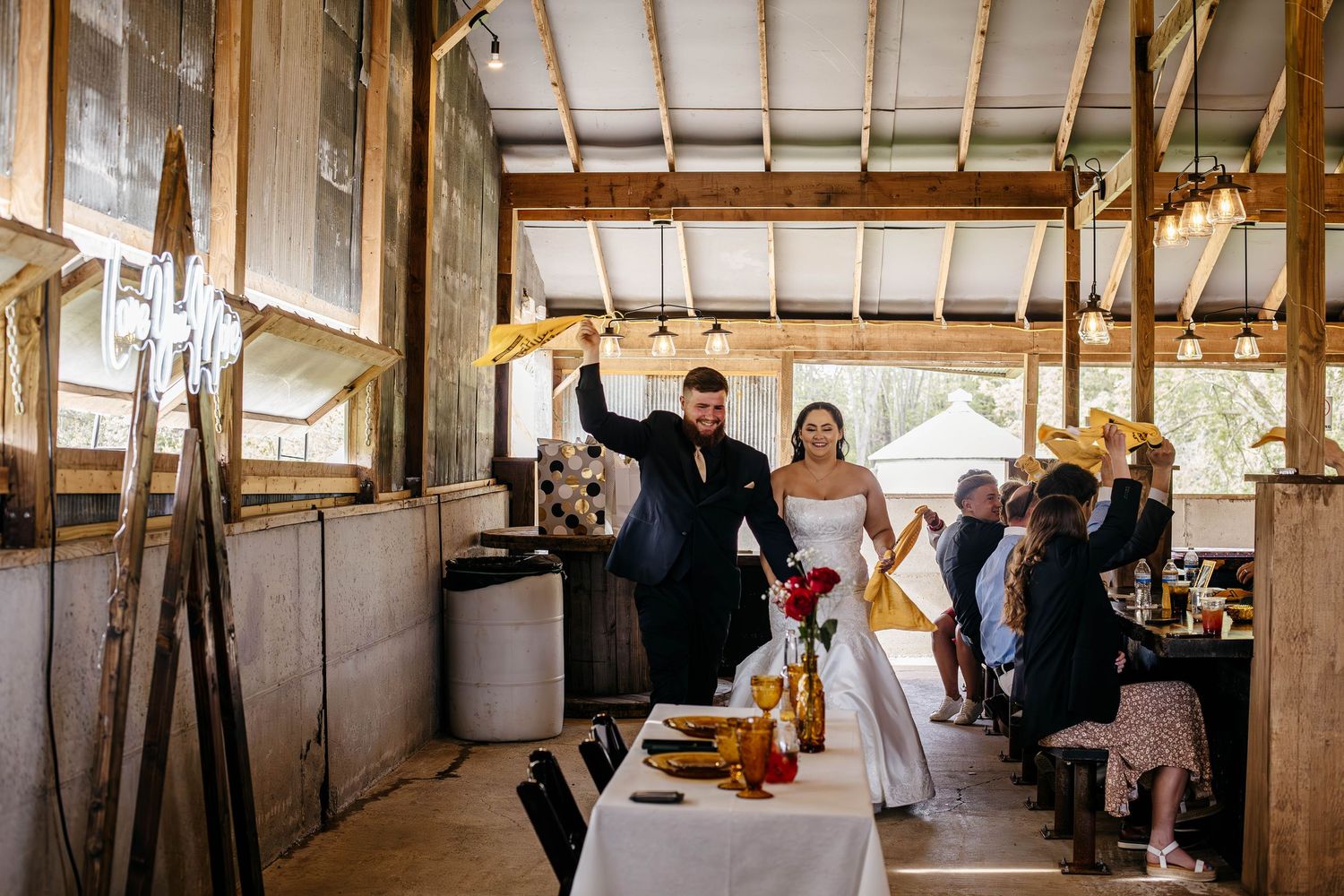 Newlywed couple celebrates with raised arms during reception in rustic barn venue surrounded by guests.