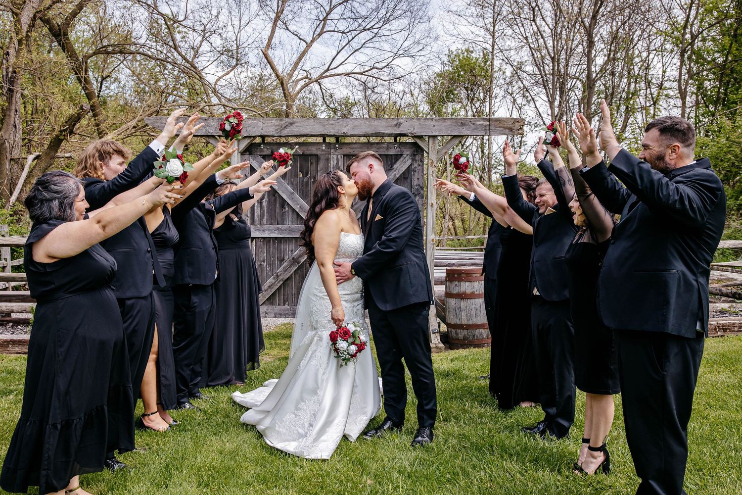Wedding party in black attire forms line outdoors with couple kissing in center during funny dabbing pose.