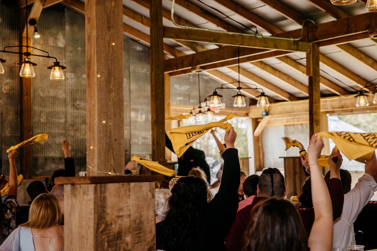 Wedding guests raise their glasses in a rustic barn venue with exposed wooden beams and warm lighting during a toast.