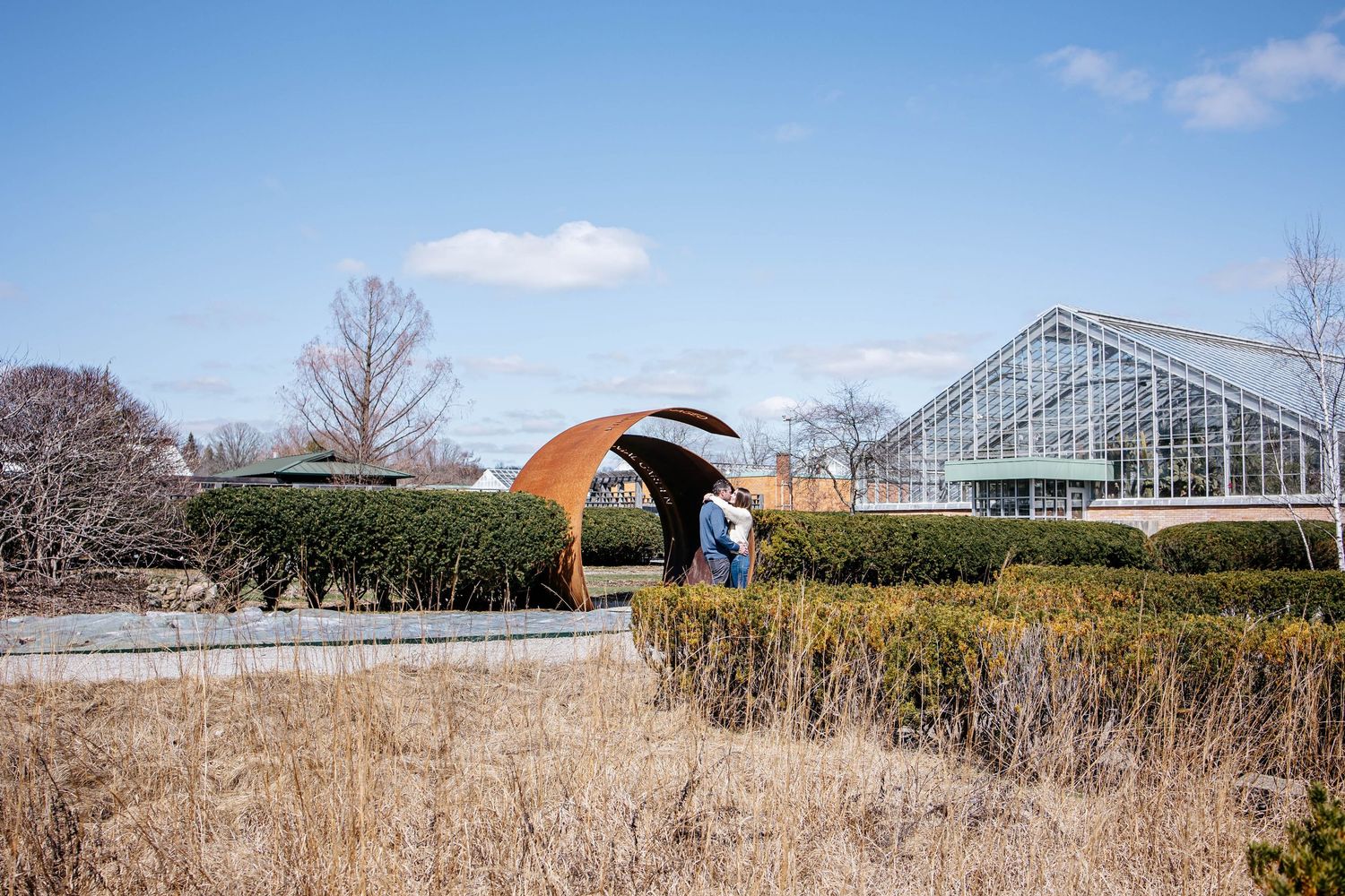 A greenhouse and sculptural archway structure stand among winter gardens at a botanical garden on a sunny day.