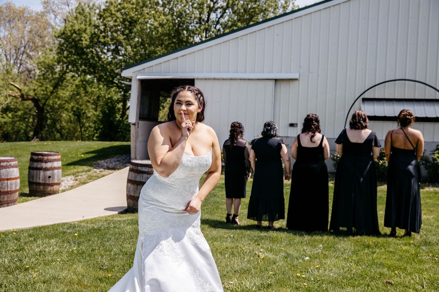 A bride in a white mermaid dress poses while attendants in black dresses stand in a line by a barn.