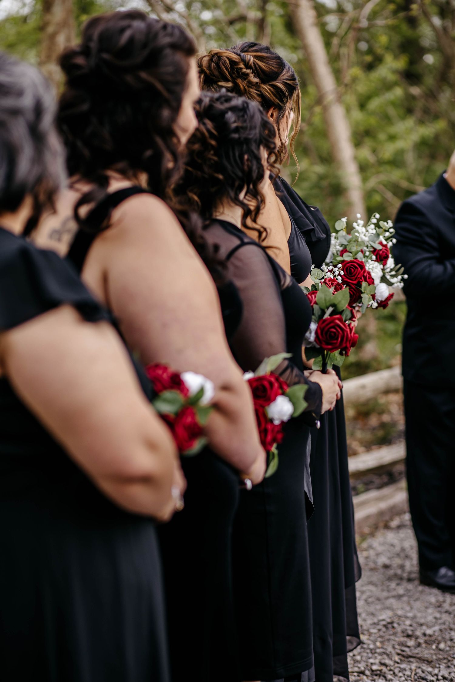A row of bridesmaids in black dresses hold bouquets of burgundy and white flowers during an outdoor wedding ceremony.