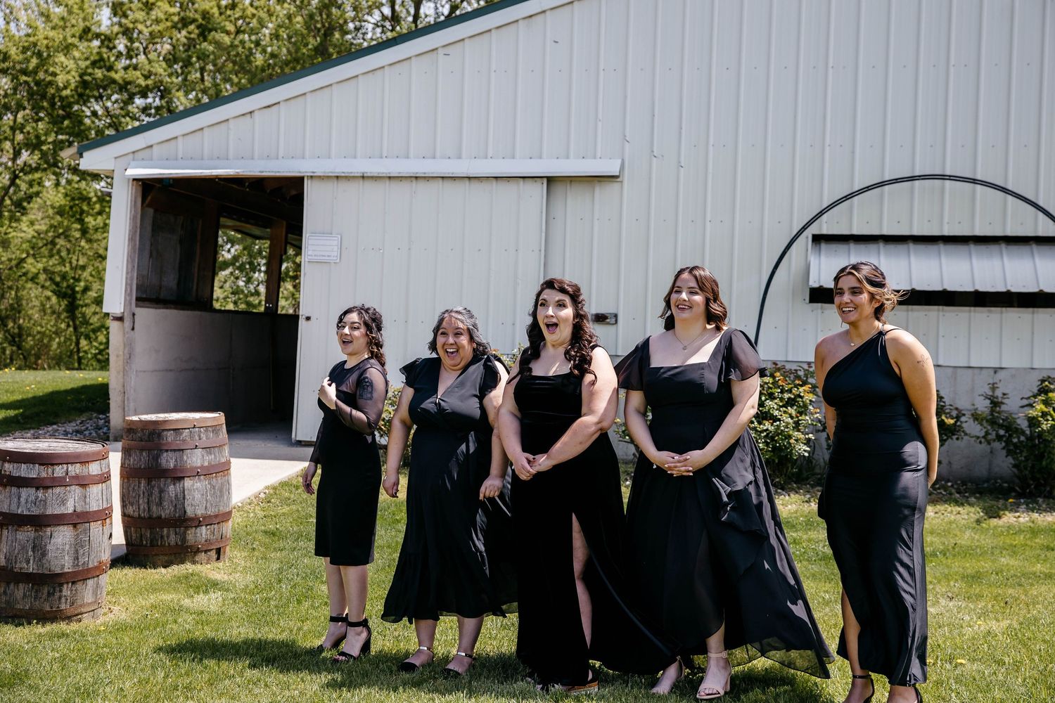 A wedding party wearing black dresses stands together outside creating lively and fun candid moments by a barn.