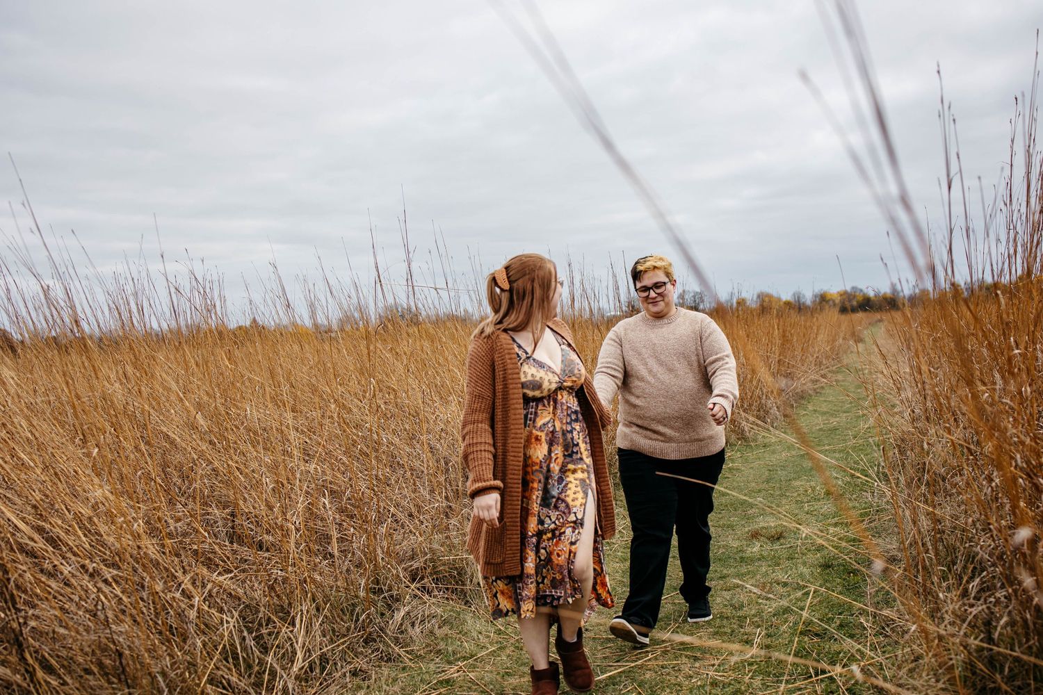 Two people walk together on a nature path through tall brown grasses on an overcast autumn day.