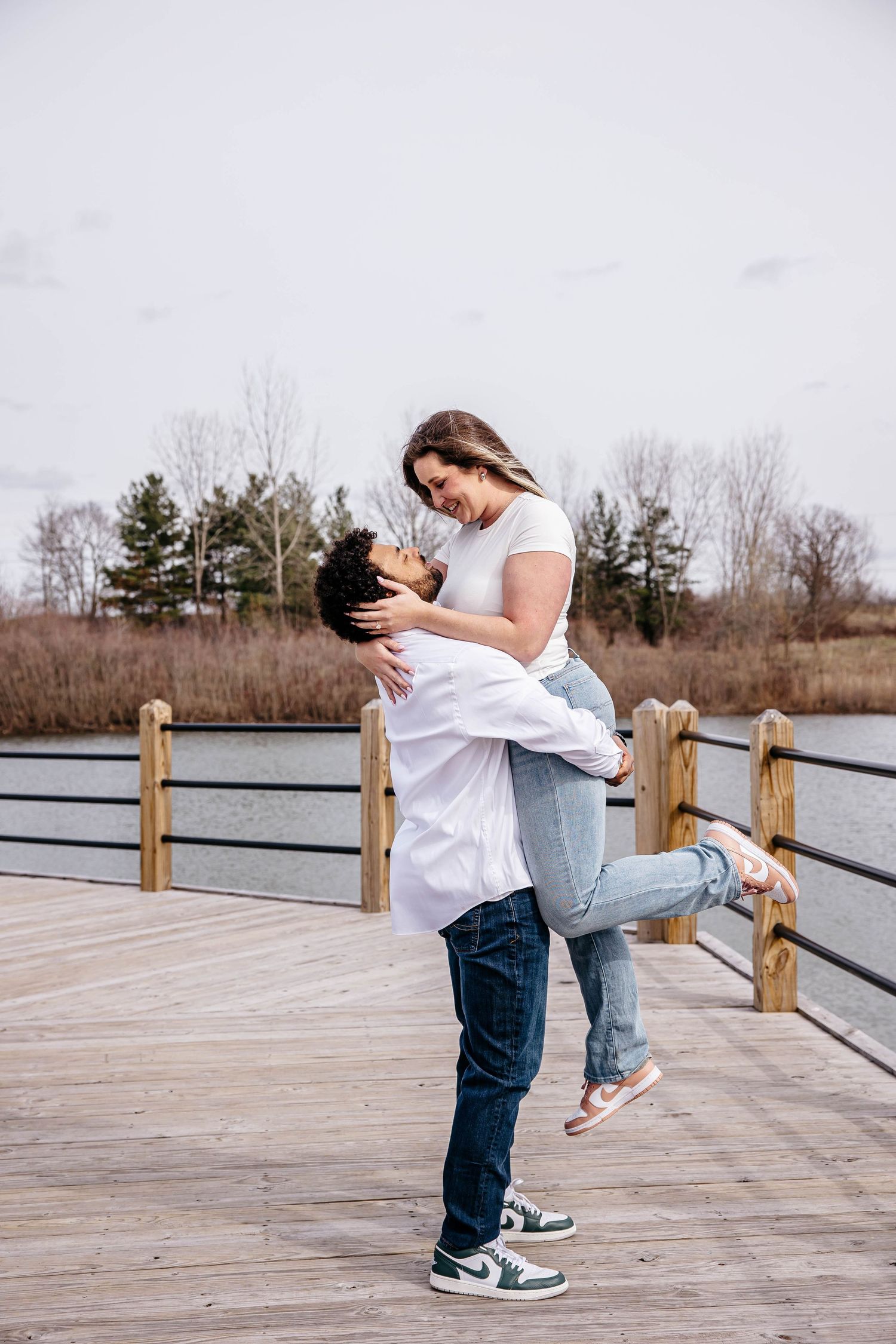 A happy couple sharing an affectionate embrace and lifting moment on a wooden boardwalk near a lake during sunset.