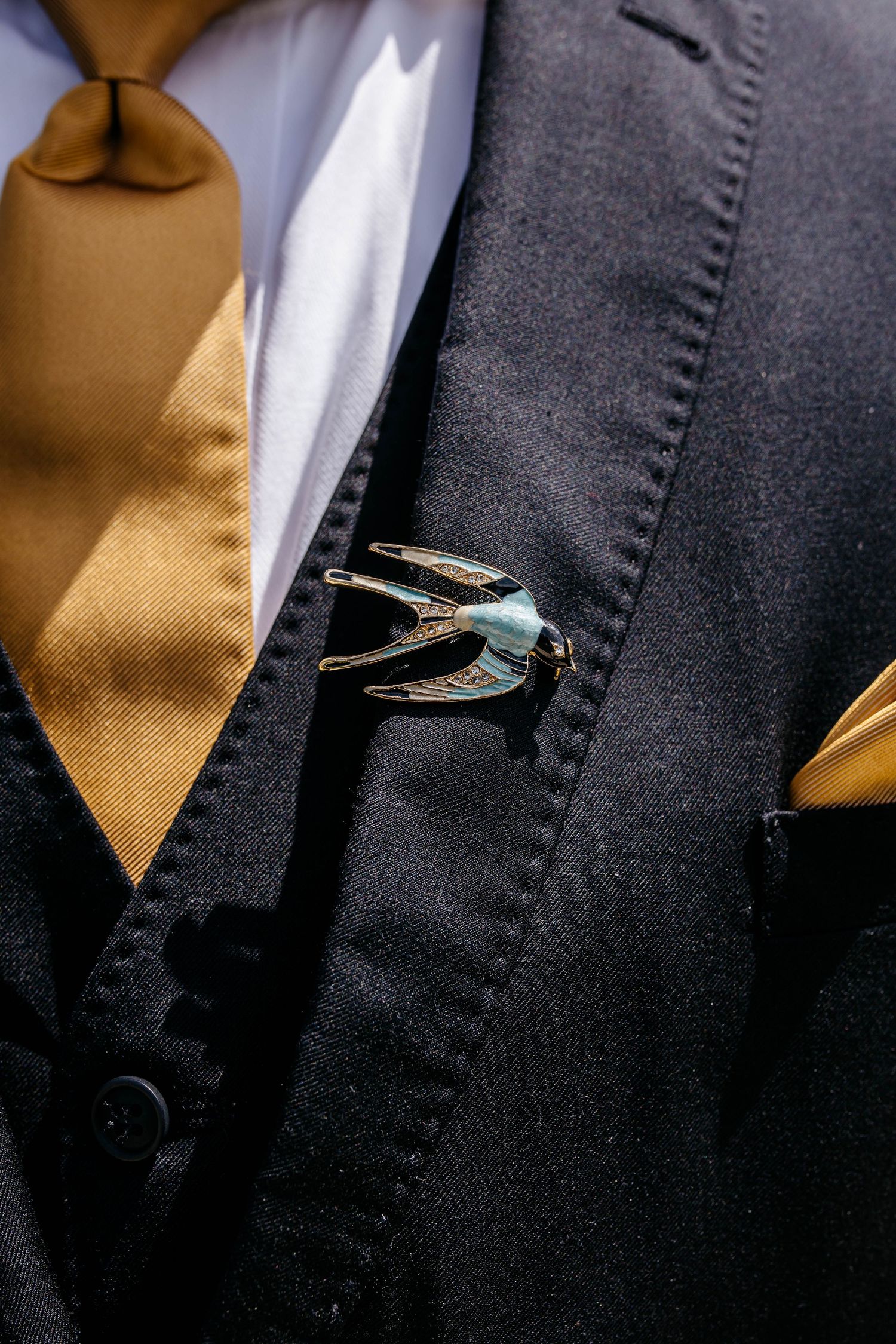 A silver swallow-shaped brooch pin adorns a dark suit vest with a golden tie peeking through.