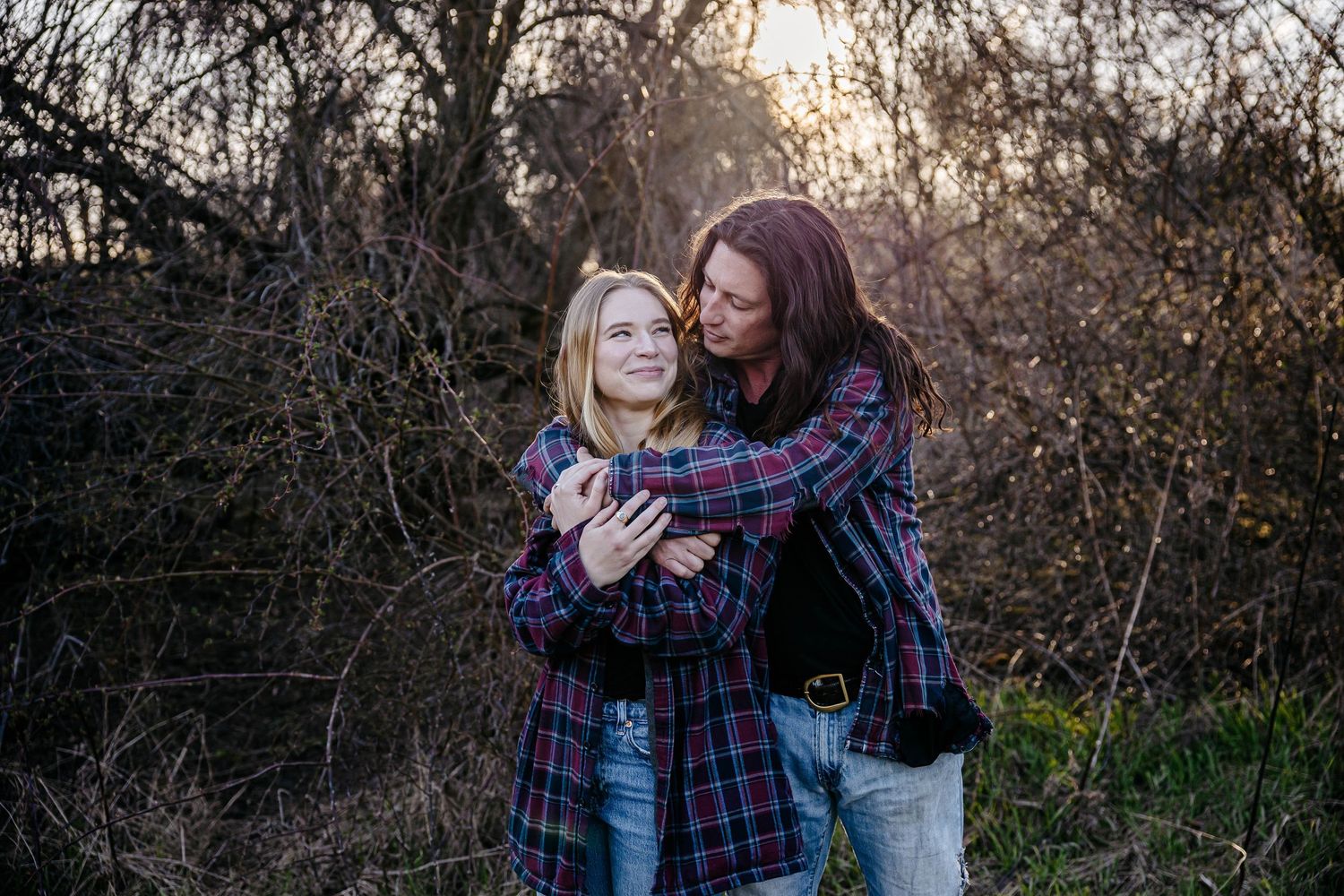 Two people embrace and laugh together outdoors at sunset wearing plaid shirts and jeans.
