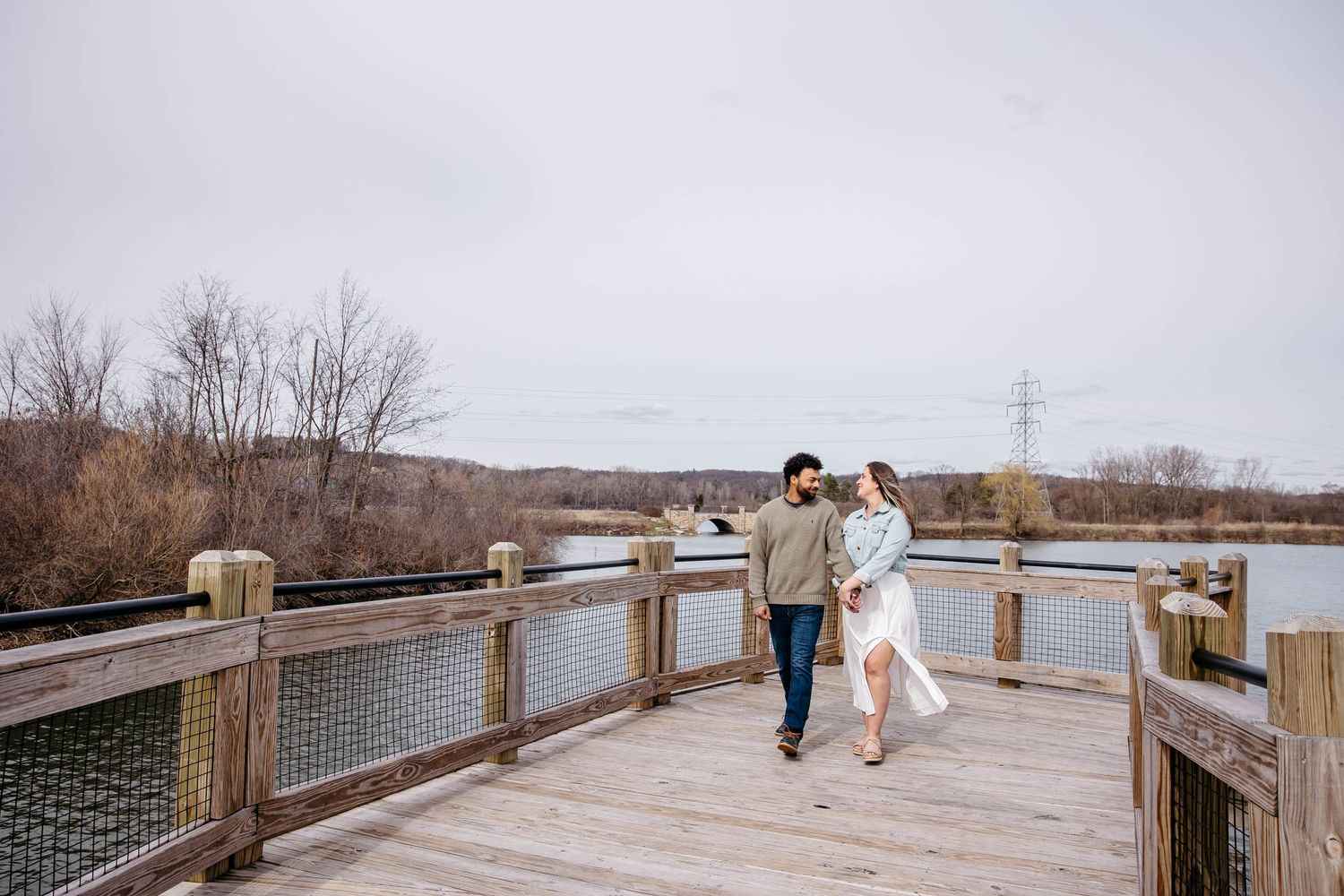Couple walking together on a wooden boardwalk over wetlands during golden hour with electrical towers in the distance.