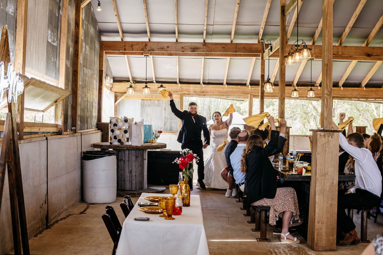 Wedding guests enjoying a rustic barn reception while servers prepare food at a long dining table in a wooden venue.