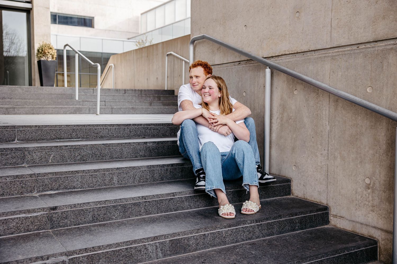 Two people sitting and laughing together on concrete steps outside a building with metal handrails.