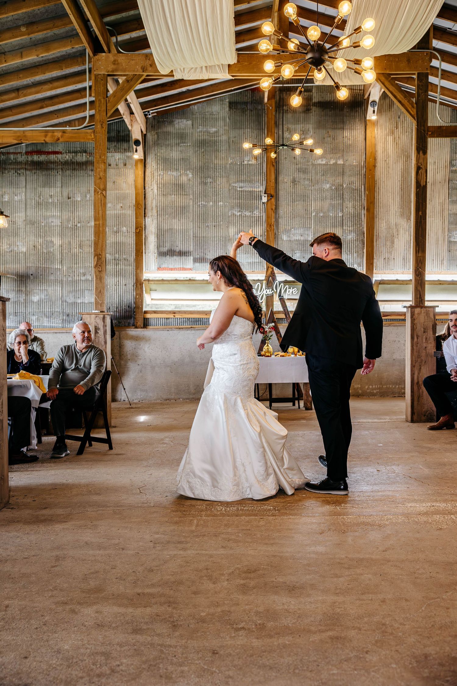 A couple shares their first dance at a rustic barn wedding venue decorated with elegant lighting and white draping.