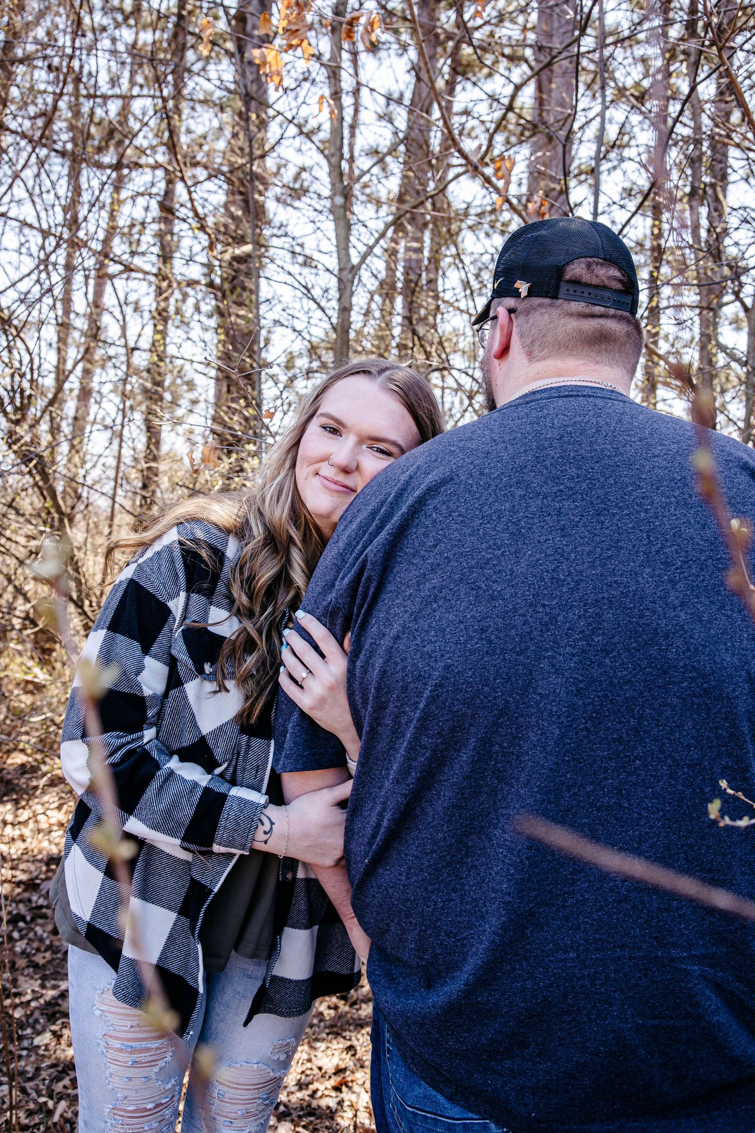 A couple embraces in an autumn forest setting with fallen leaves and bare trees in the background.