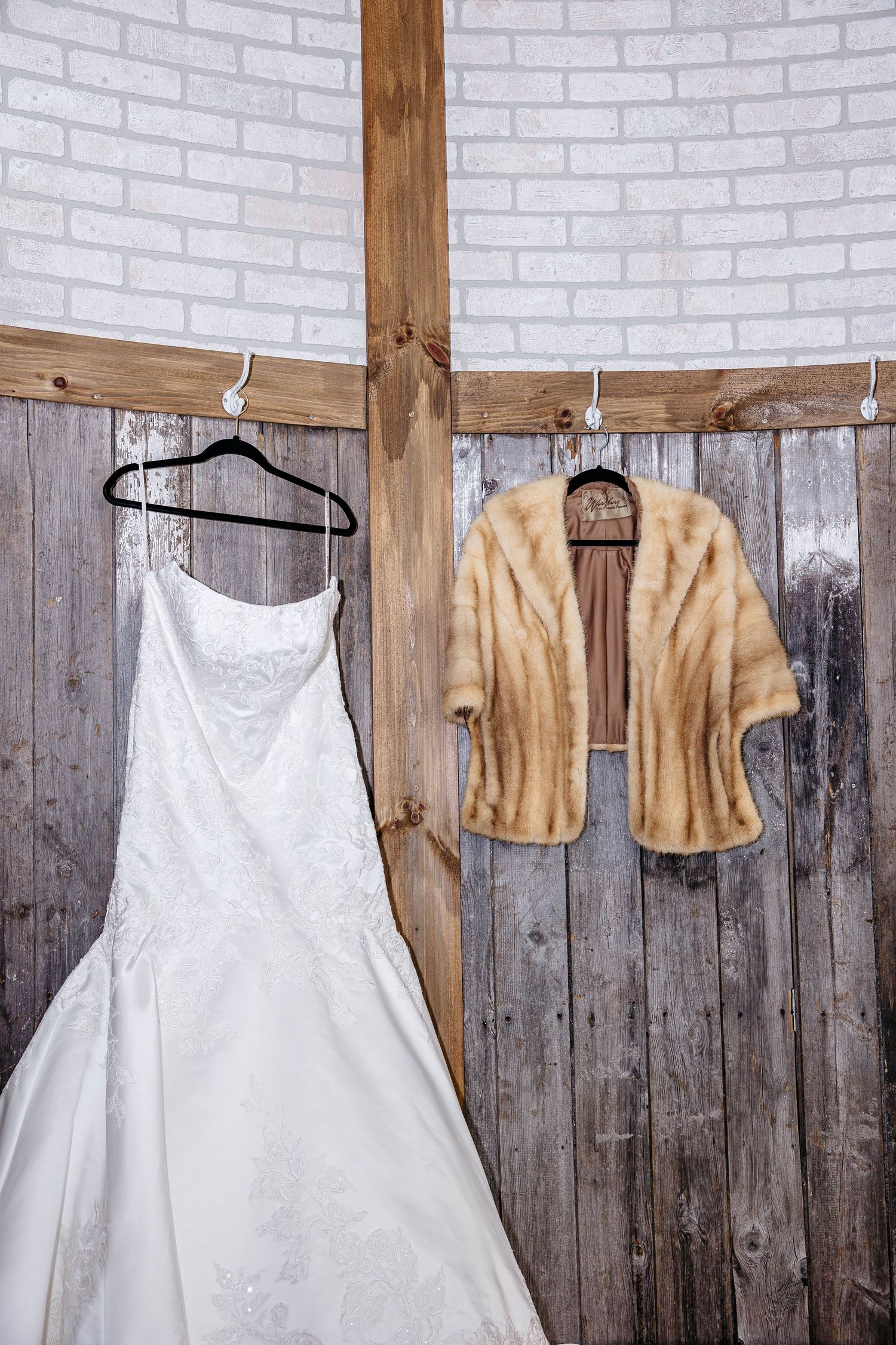 A white wedding dress and vintage fur stole hang on wooden hangers against a rustic brick and wood wall background.