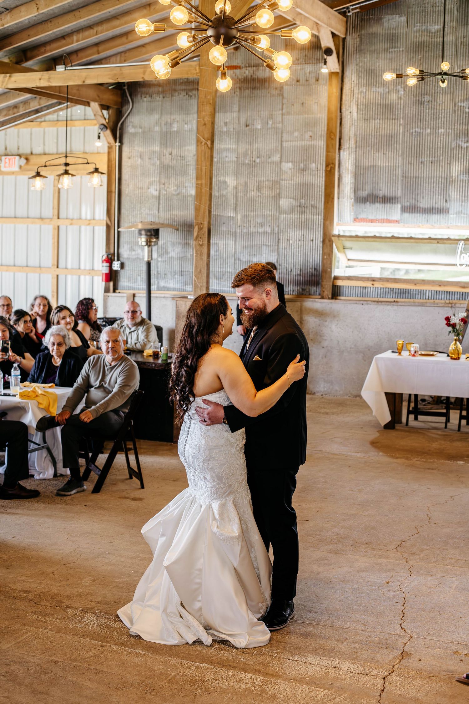 A couple shares their first dance in an industrial-style wedding venue with exposed walls and modern chandeliers.