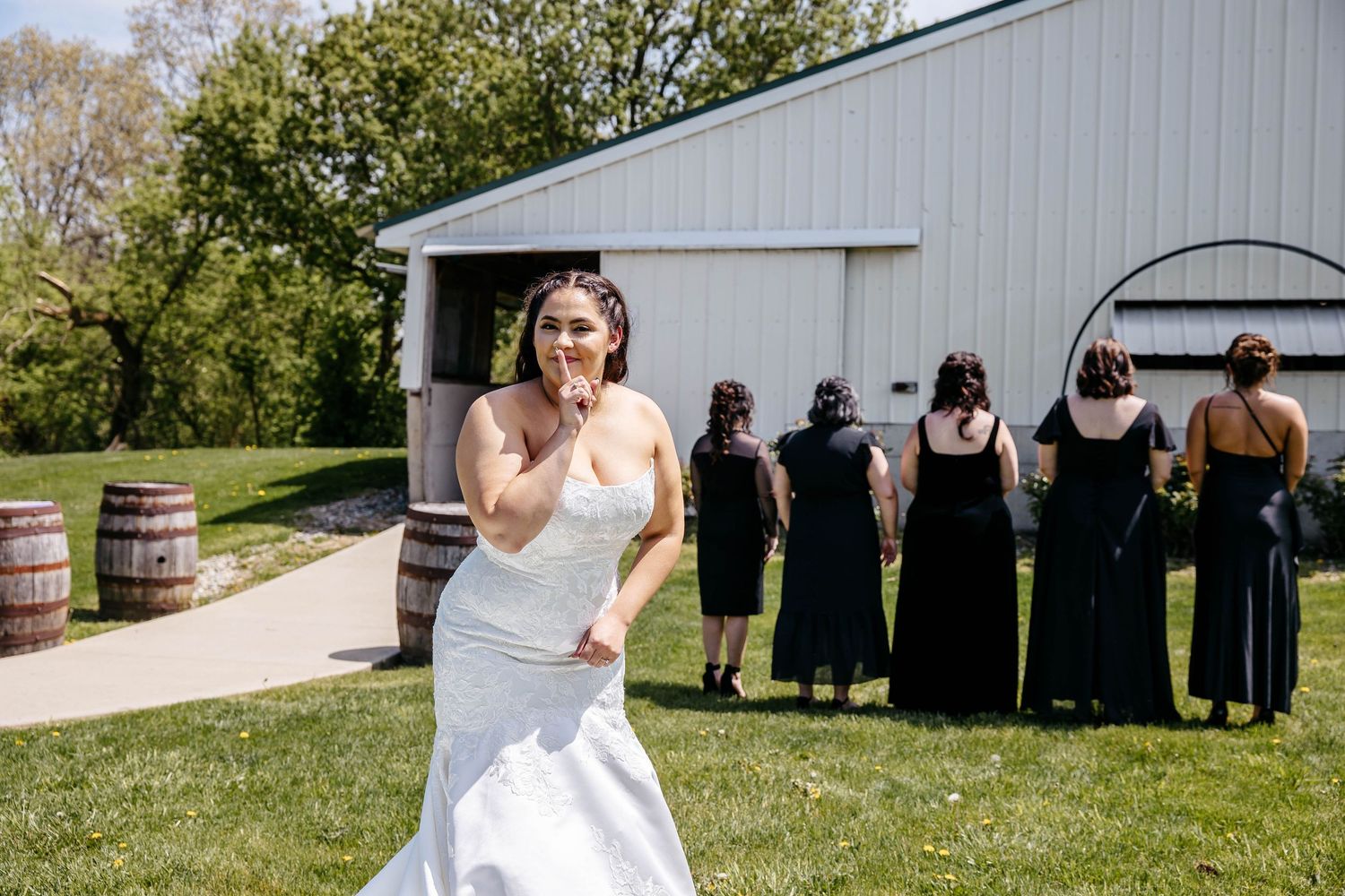 A group of women in black dresses stand behind a white barn while a bride poses in front on a sunny day.