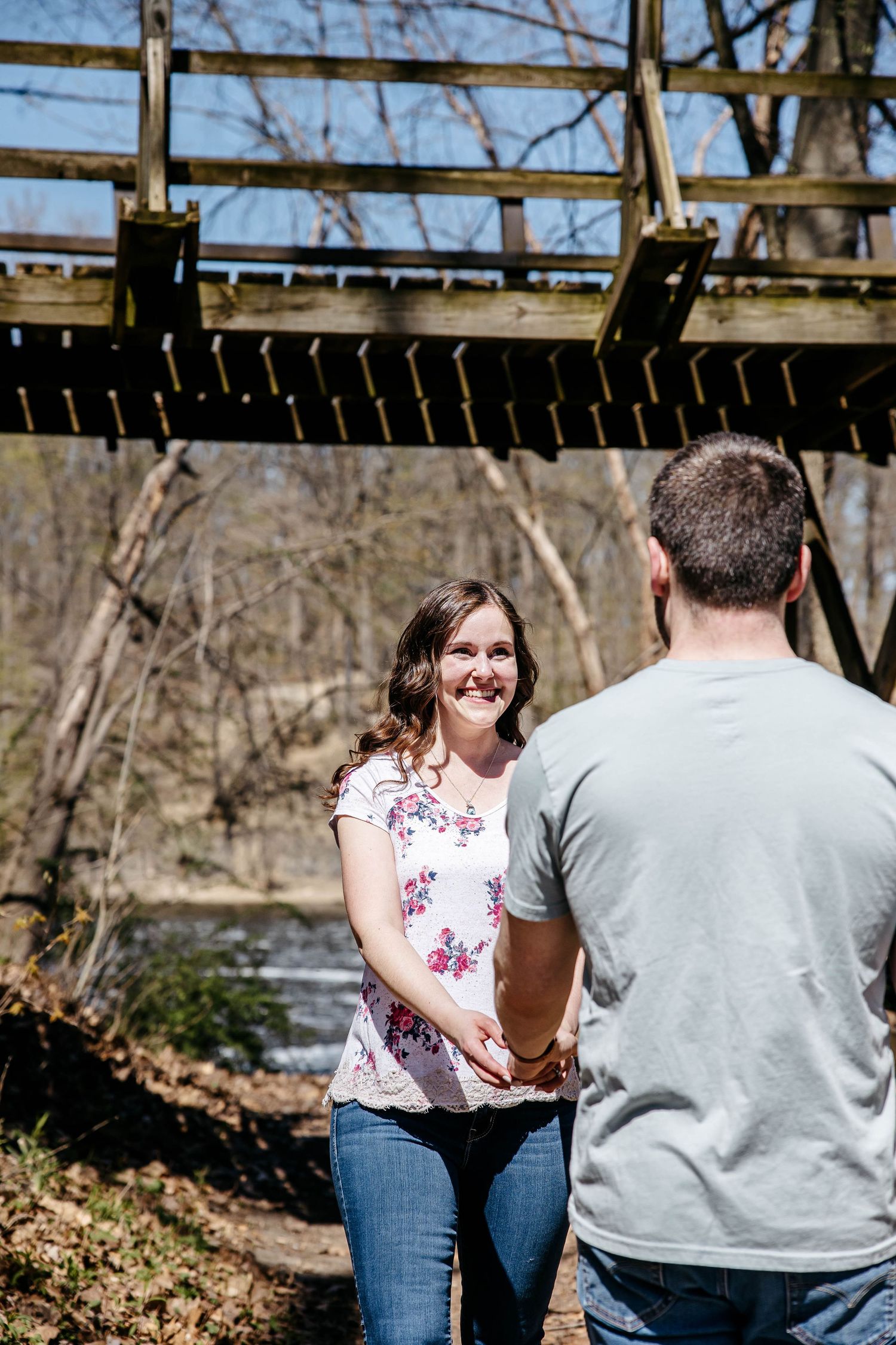 A couple shares a joyful moment together beneath a rustic wooden bridge on a sunny spring day.