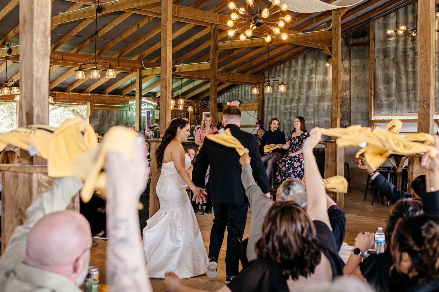 Wedding guests dance and celebrate in a rustic barn venue with wooden beams and string lighting overhead.