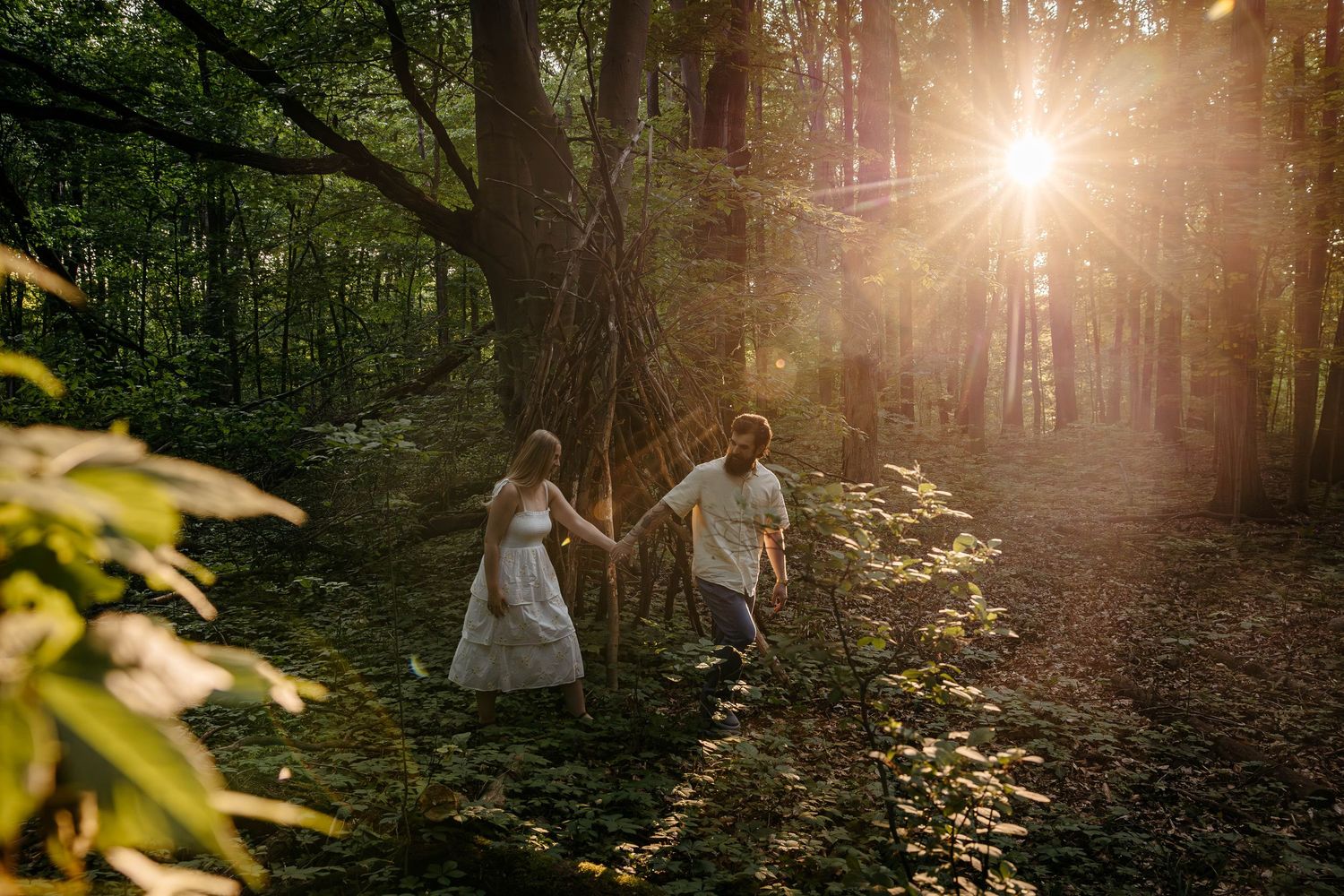 Two people walk hand in hand through sunlit forest path during golden hour as sun rays stream through trees.