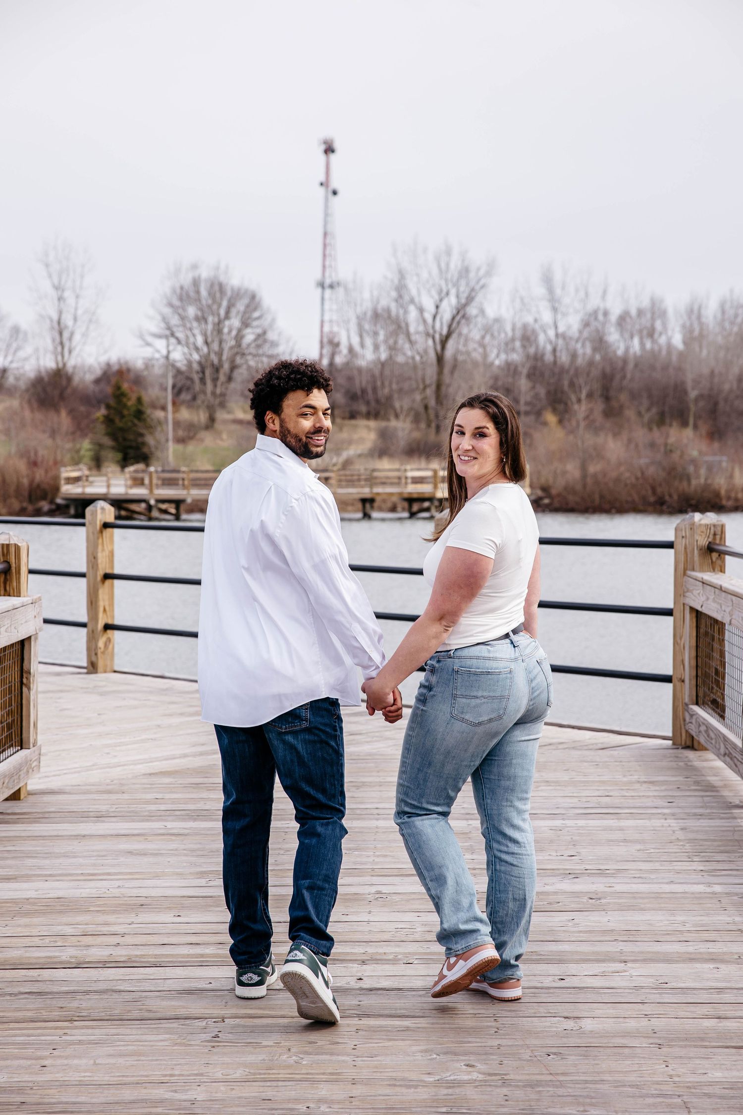 A couple shares playful moments while walking on a wooden boardwalk by a lake during an engagement photoshoot.