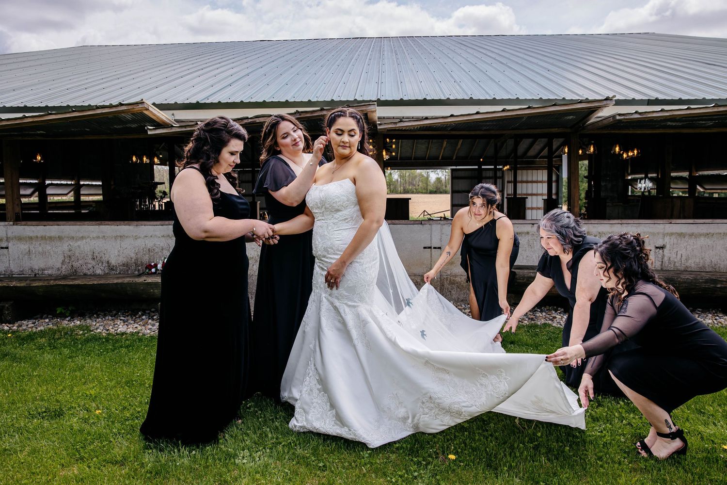 Wedding party gathers outdoors by a rustic barn venue, with bridesmaids in black helping adjust a flowing white gown.