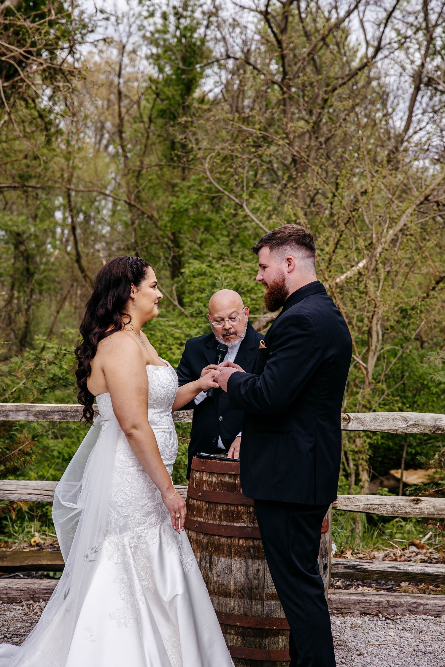 Couple during their outdoor wedding ceremony beside a rustic wooden barrel and fence in a wooded setting.