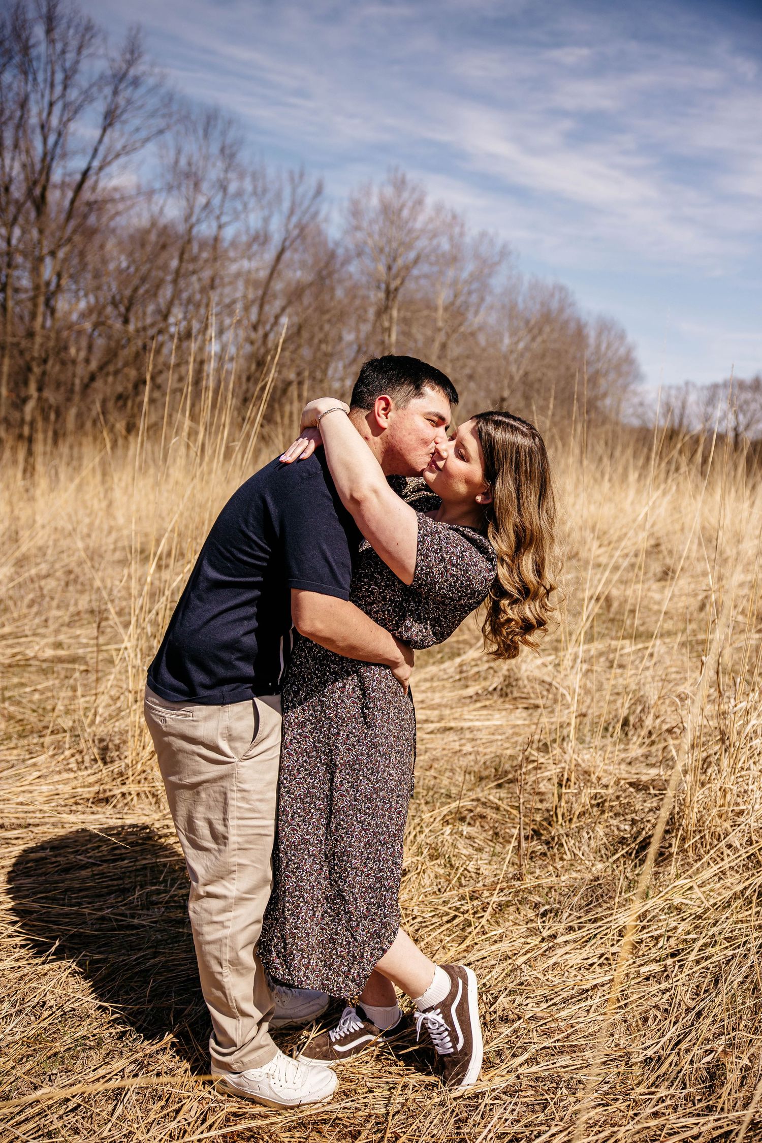 A romantic couple embraces and kisses in a golden field of tall grass on a partly cloudy day.