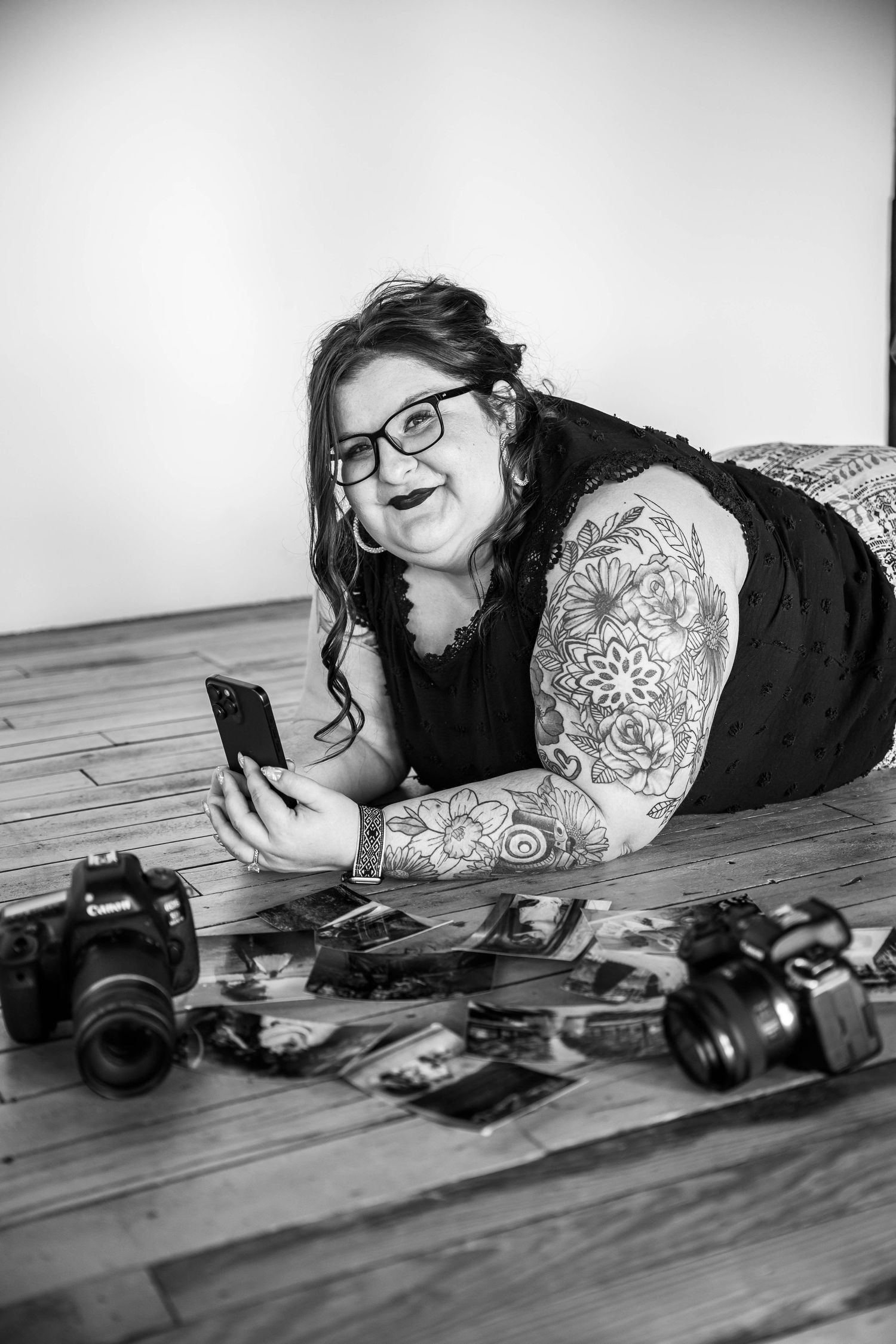 Black and white photo of a photographer on floor surrounded by cameras, showing an arm with floral tattoos looking at phone.