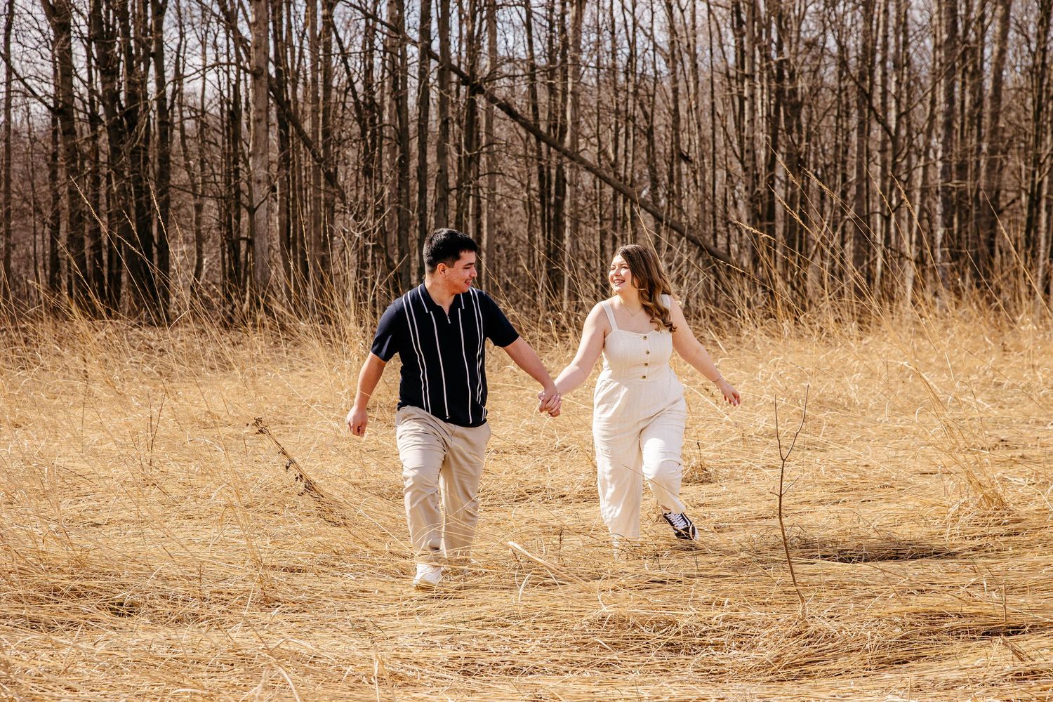A couple in coordinated black and white outfits runs joyfully through a golden field with bare trees in the background.