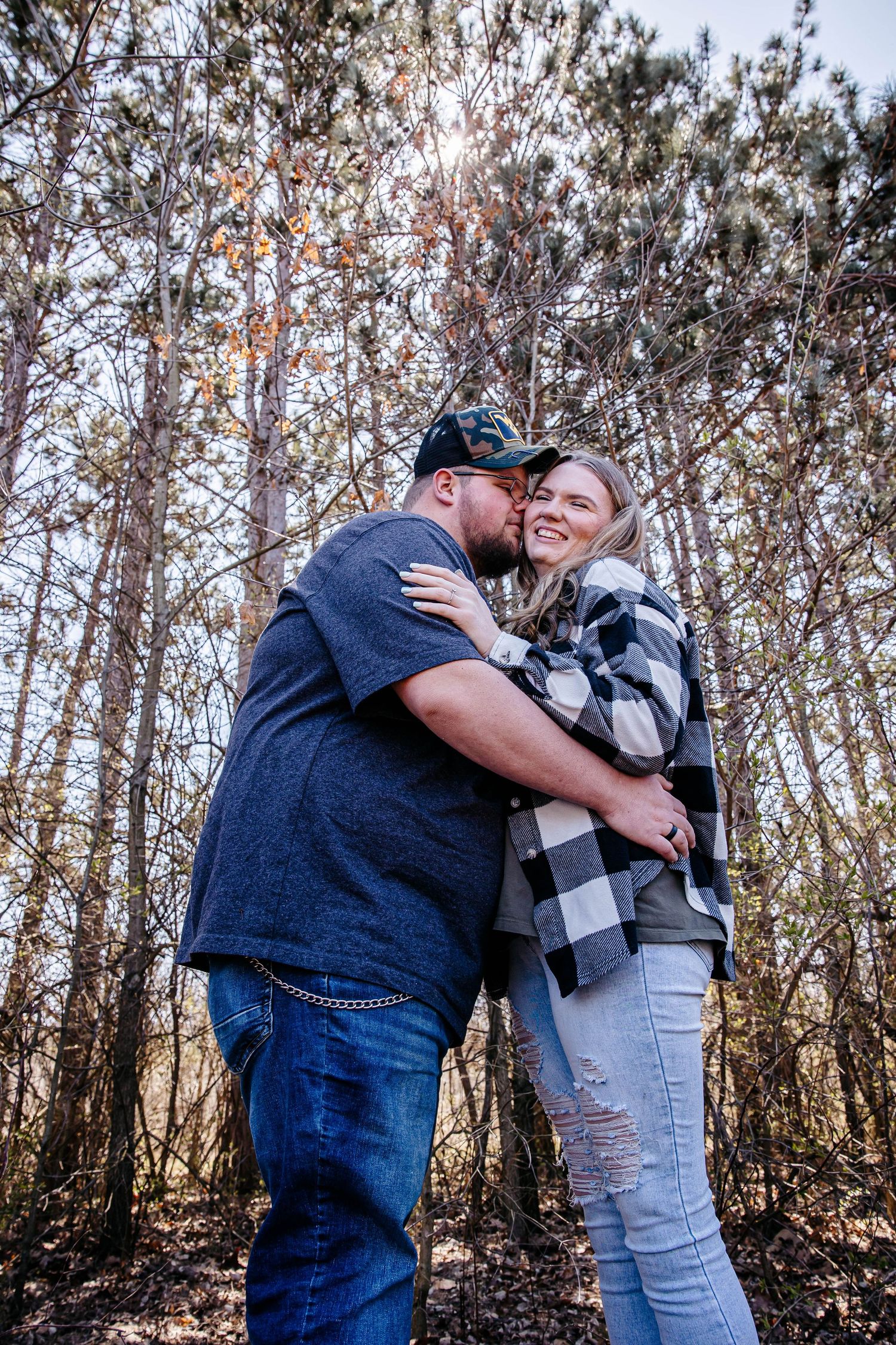 A couple share an intimate embrace in a wooded forest setting wearing casual winter clothing.