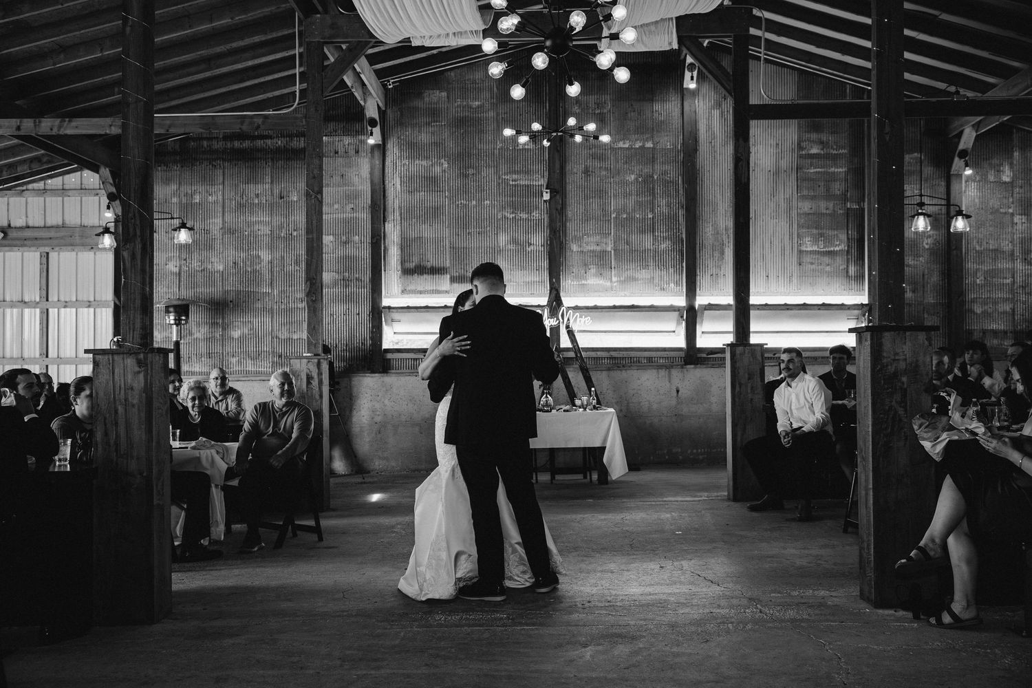 A couple shares their first dance as newlyweds in a rustic industrial venue with hanging lights creating a romantic ambiance.