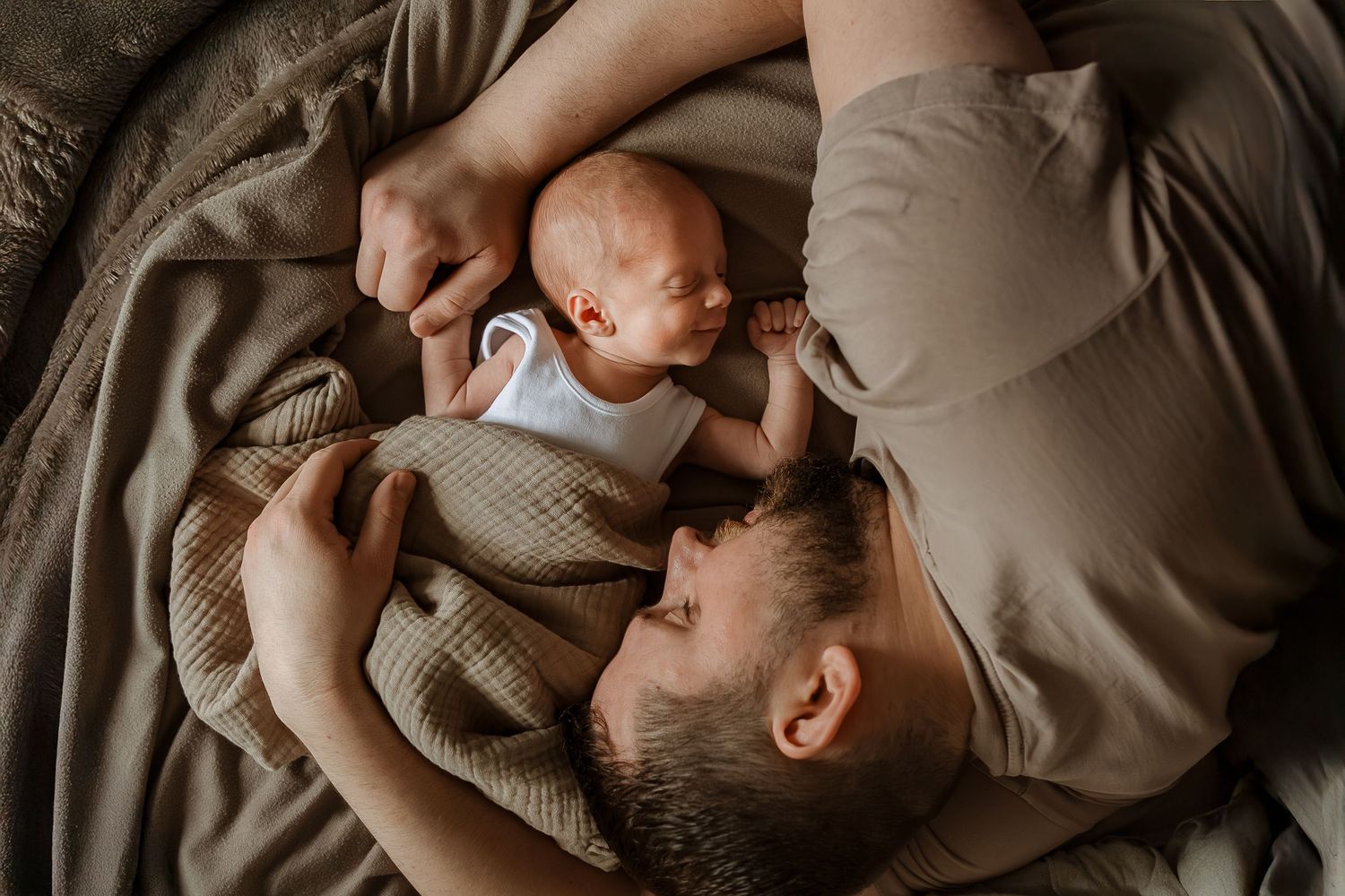 A tender moment captured from above as a parent lies on beige bedding with their newborn baby.