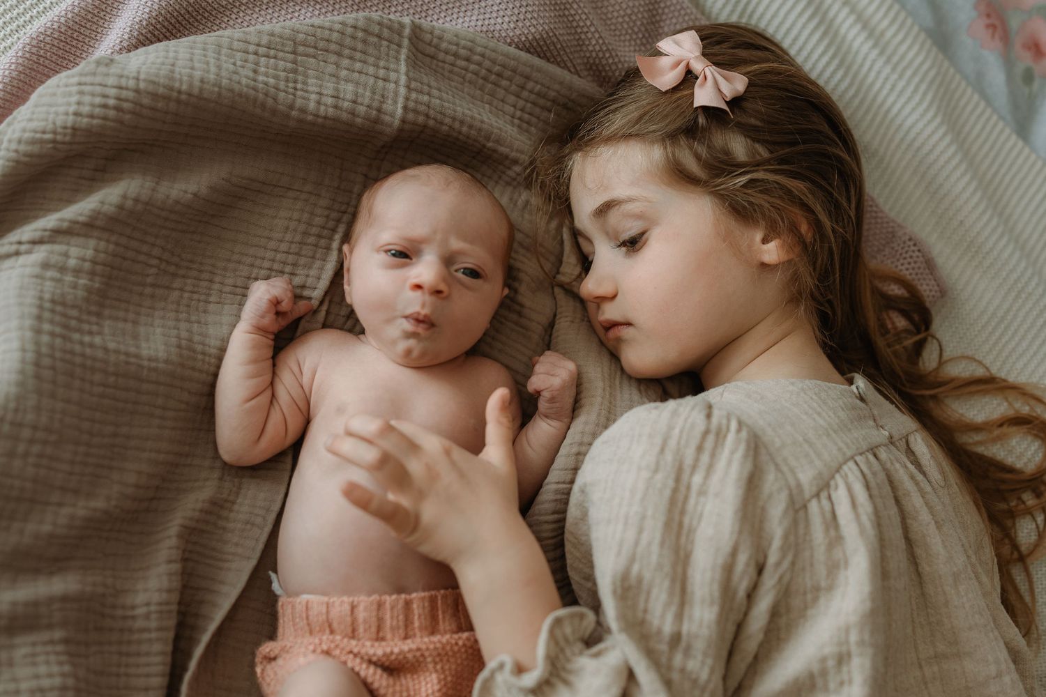 Two siblings lying together on gray bedding, with one wearing a cream-colored dress and the other in pink shorts.