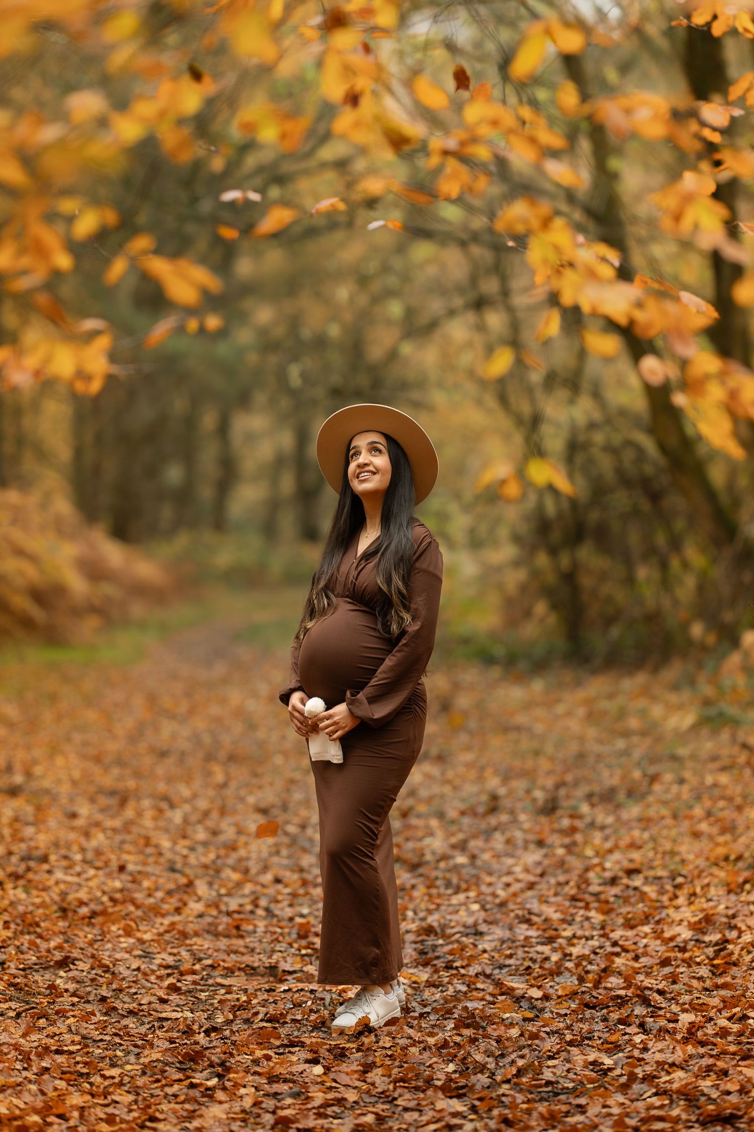 Expectant mother in brown dress and hat stands on autumn path covered in fallen orange leaves.