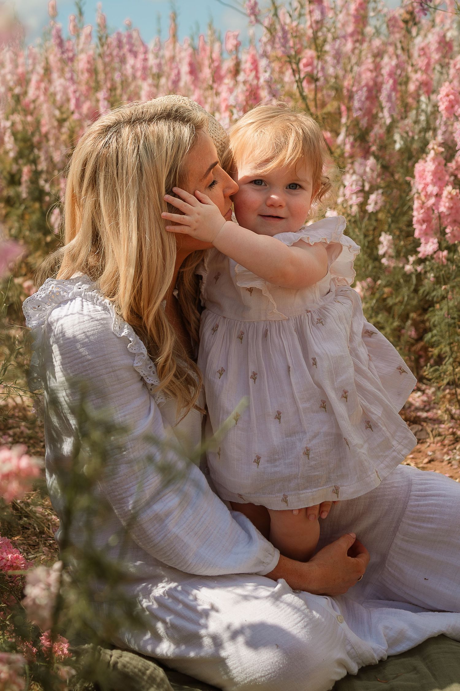 A tender moment captured in a garden with pink flowers as two people in white dresses embrace in soft sunlight.