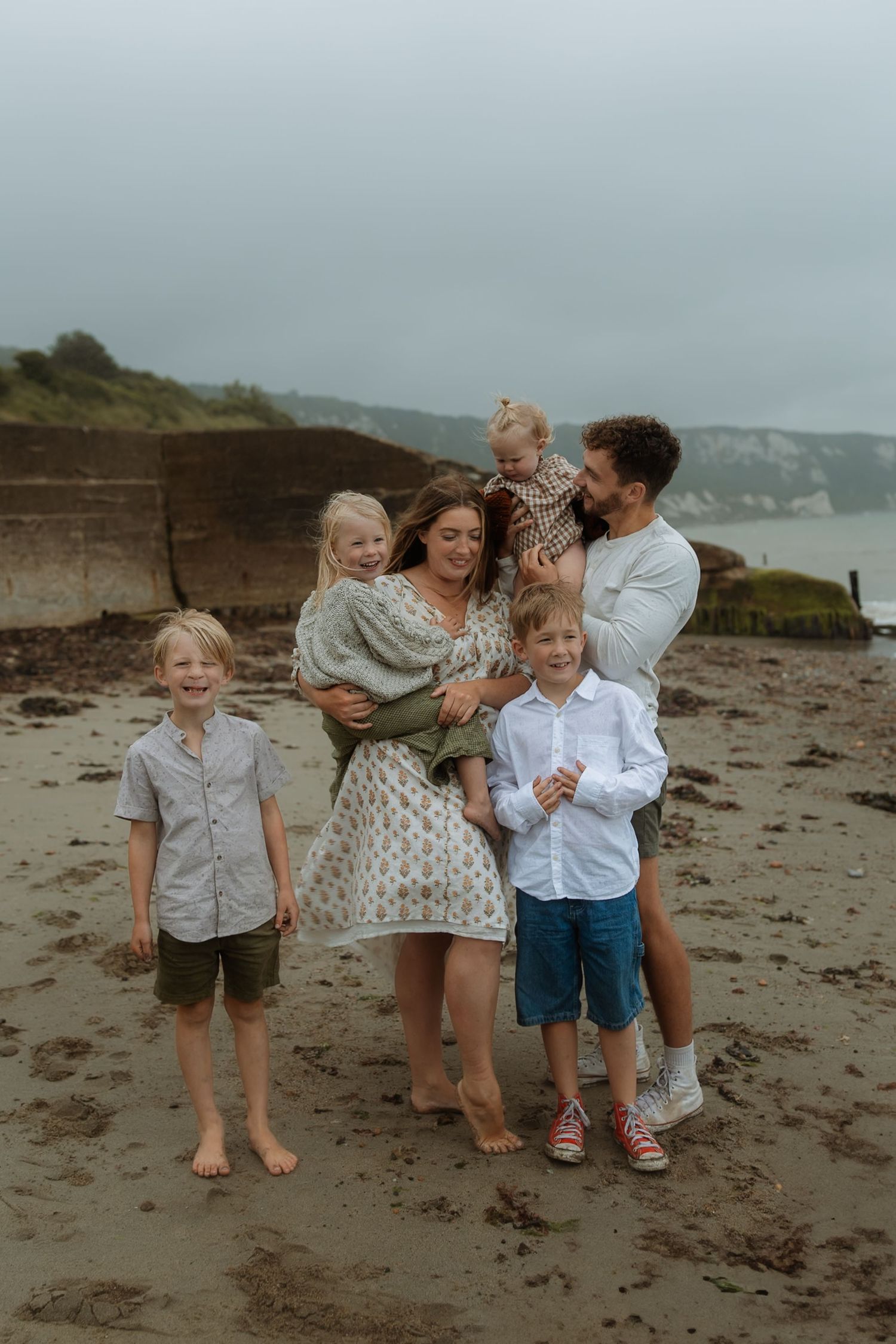 Family enjoying a playful moment together on a cloudy day at a rocky beach with mountains in the background.