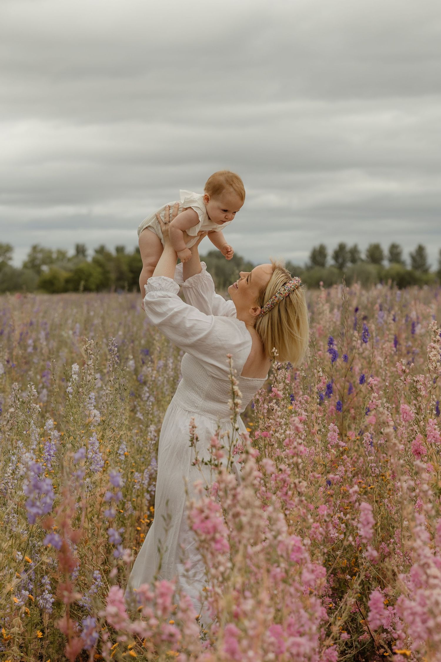 Mother in white dress lifting baby in wildflower field with pink and purple blooms under cloudy sky.
