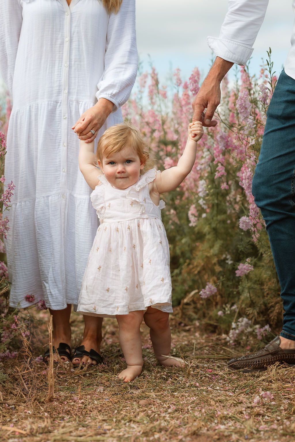 Toddler girl in white dress holds parents hands while standing barefoot in a pink wildflower field.