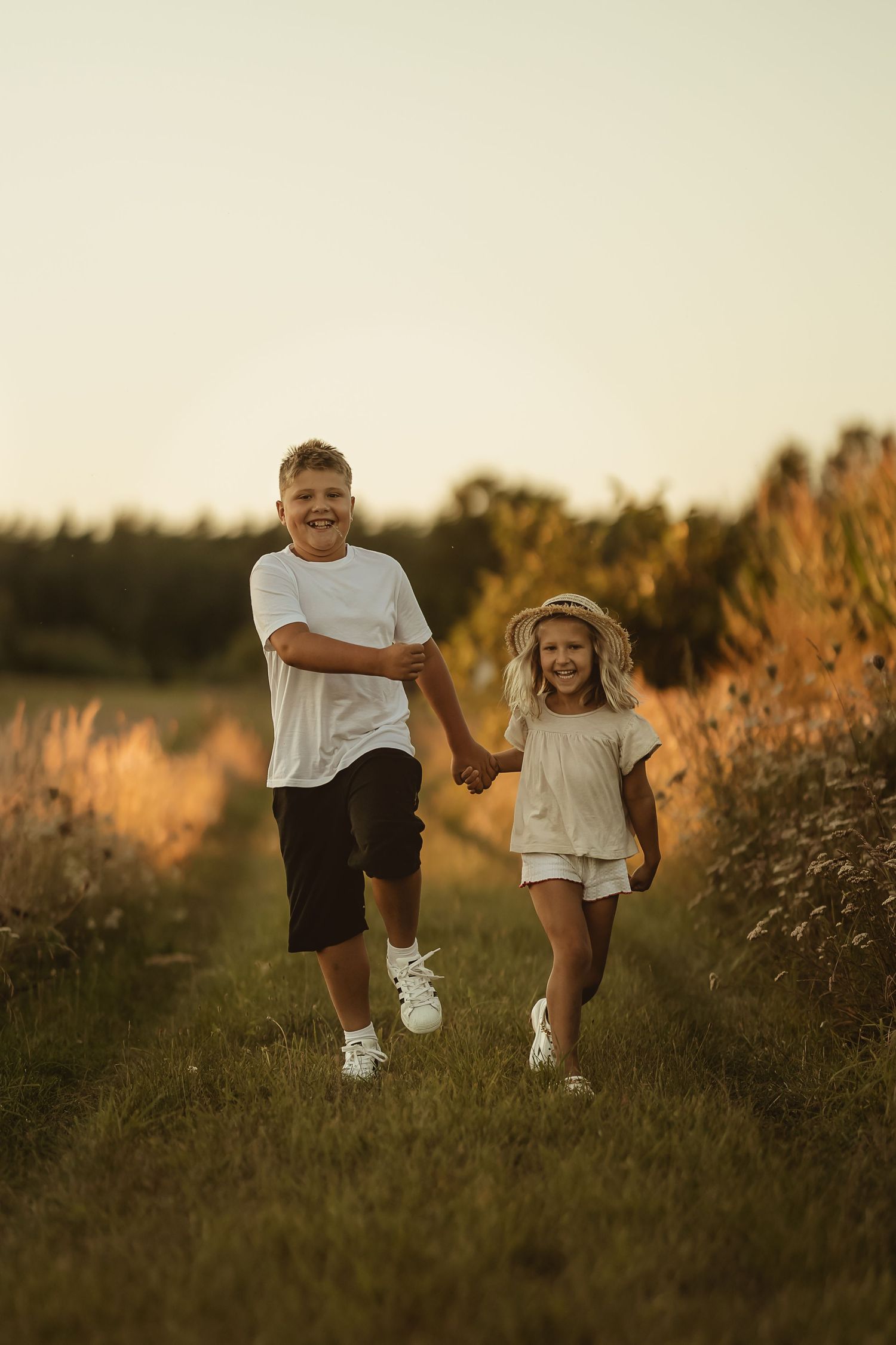 Two children run together through a golden field at sunset in matching white shirts and dark shorts.